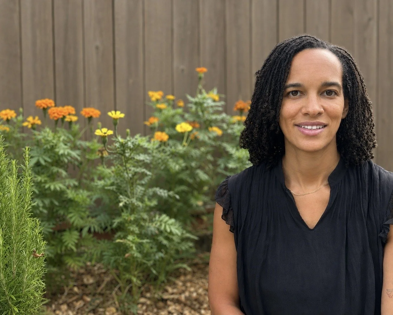 A woman with curly hair and a black top standing in a garden with orange and yellow flowers, a wooden fence in the background.