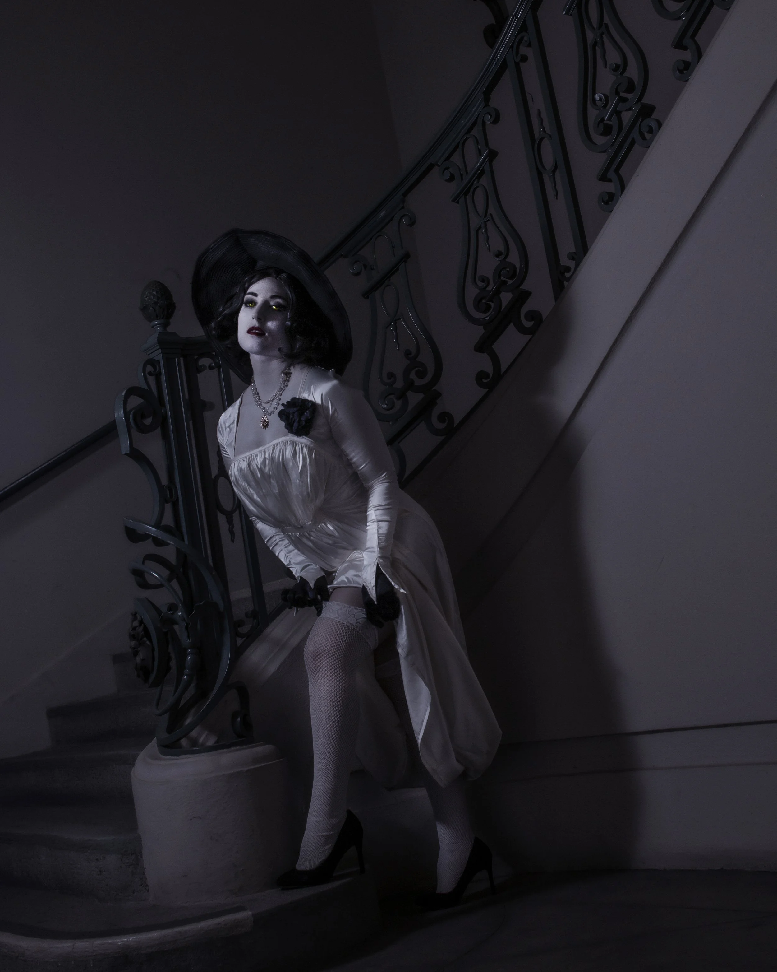 A woman dressed in vintage style clothing with a black hat, dark lipstick, and jewelry, leaning against a staircase with ornate black wrought-iron railing in a dimly lit setting.