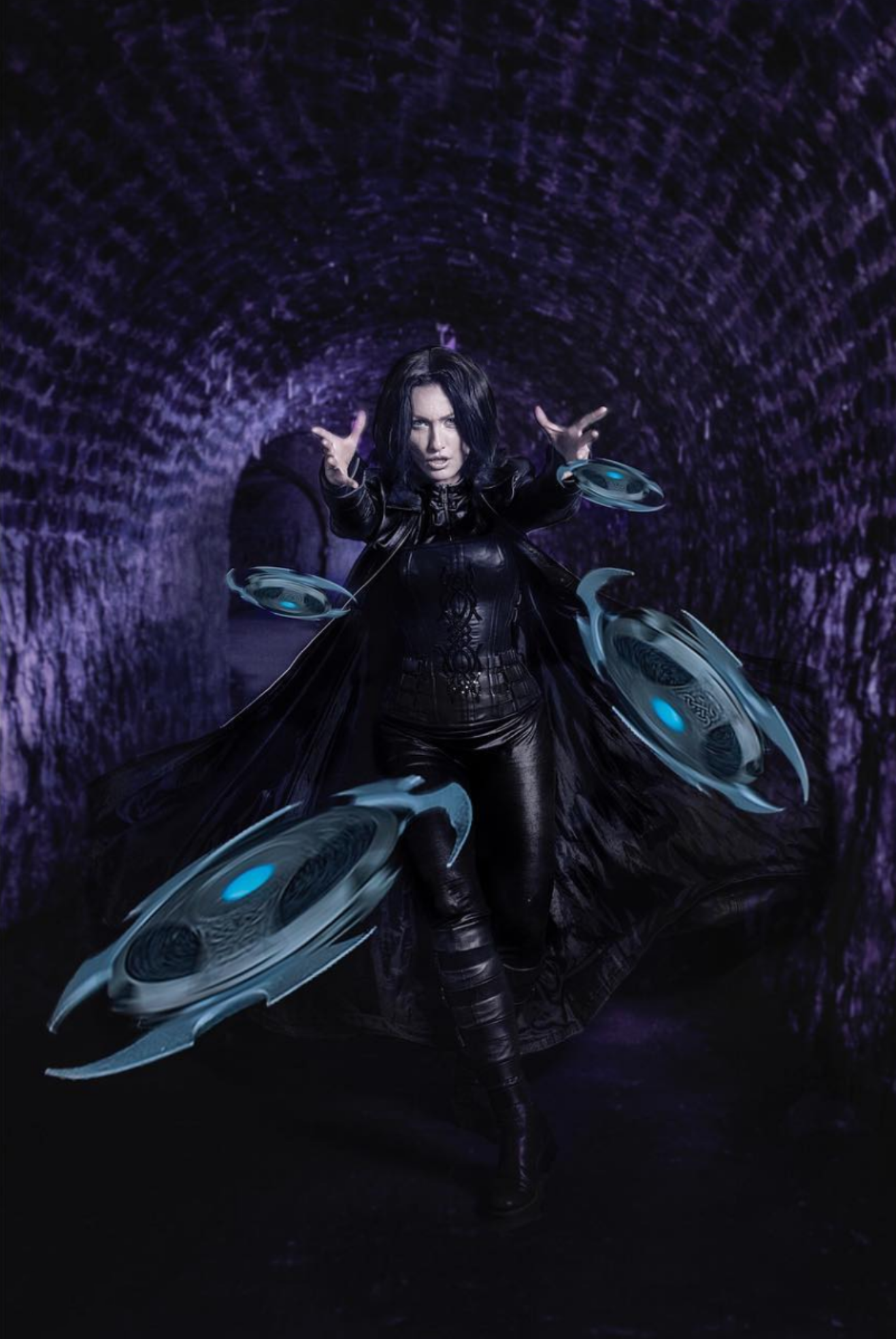 A woman dressed in black with messy black hair, standing in a dark tunnel with purple lighting. She is reaching out with her hands towards floating blue discs with black centers, creating a glowing effect around her.