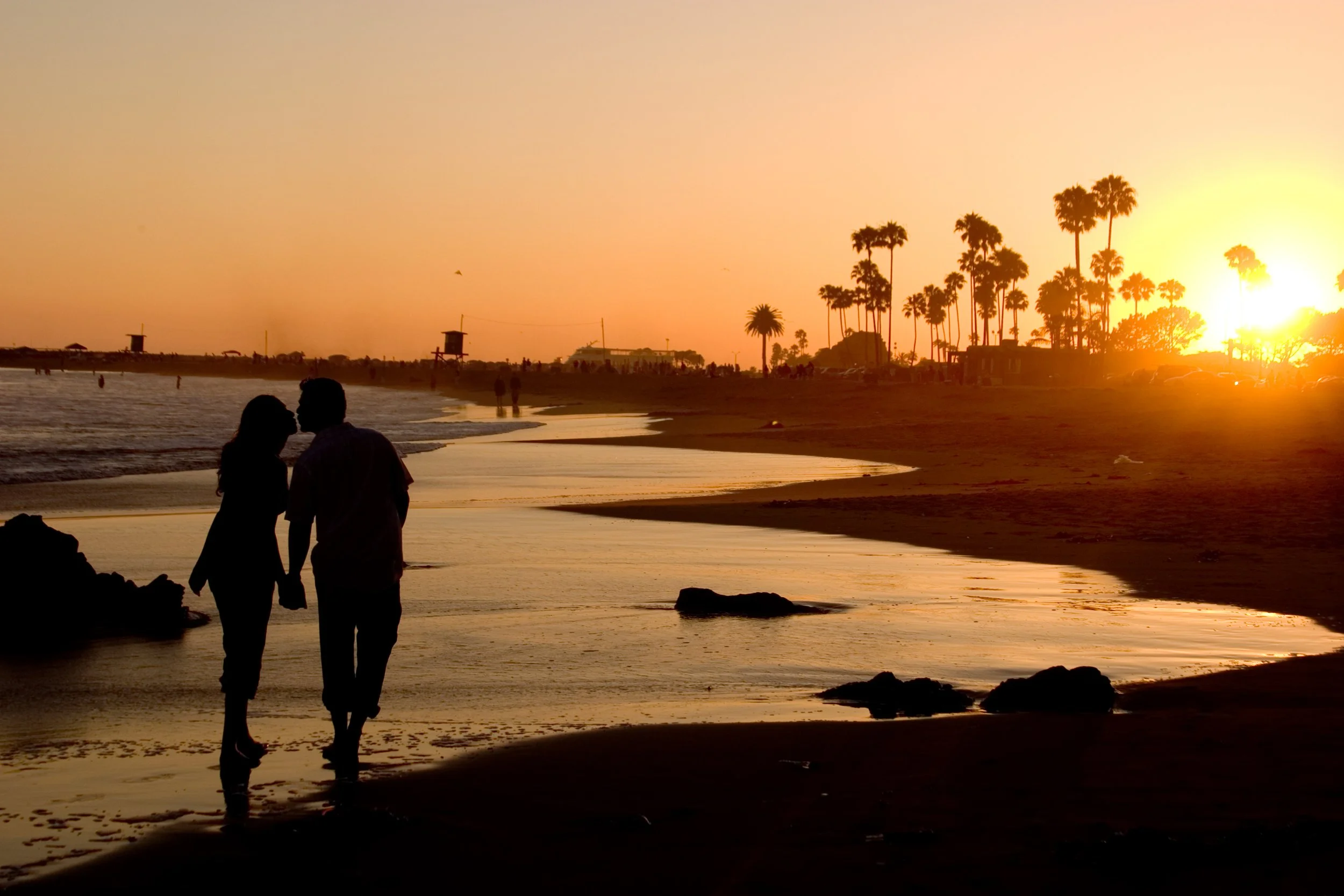 Couple walking along the beach at sunset with palm trees and lifeguard towers in the background.