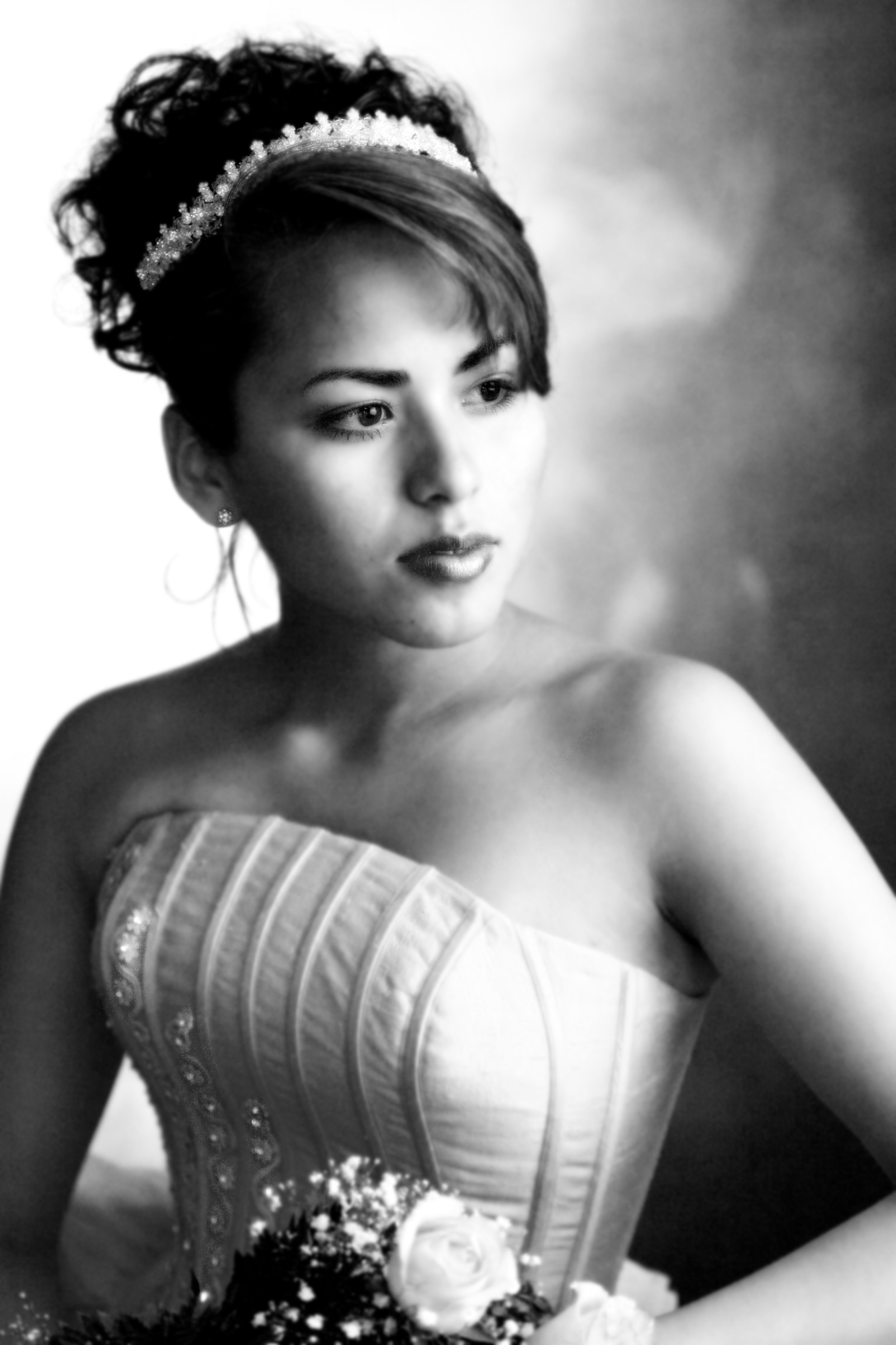 Black and white portrait of a woman with short hair styled back, wearing a strapless dress with detailed embroidery, and a floral headpiece, looking to the side, with a blurred background.