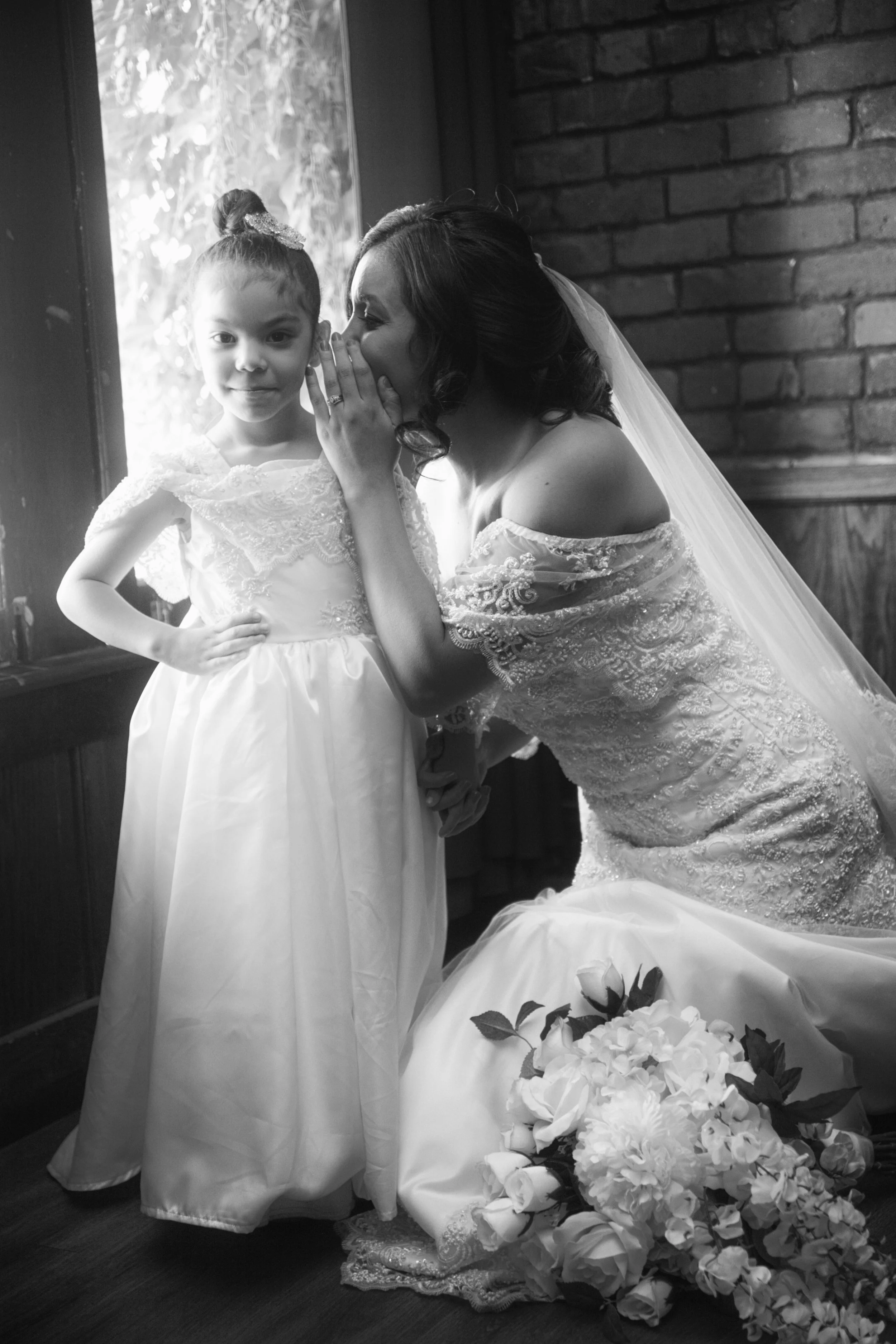 A bride whispering to a young girl in a wedding dress, with a bouquet of flowers on the floor, near a window and brick wall, in black and white