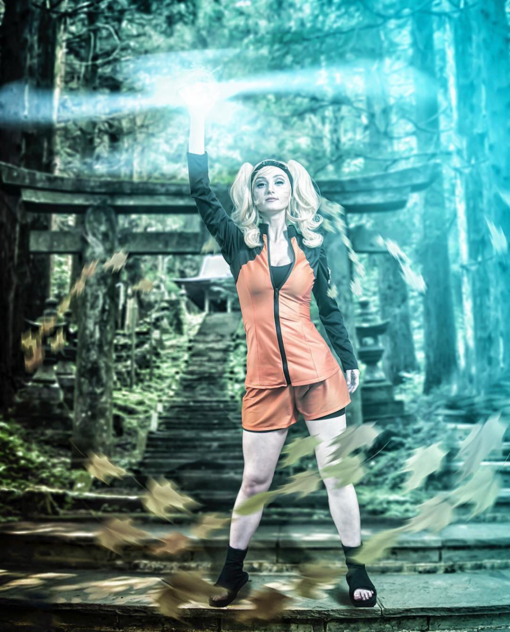 A woman with blonde hair in pigtails and light skin poses outdoors on a wooden pathway surrounded by trees. She is wearing a black and orange jacket, brown shorts, white tights, and black footwear. She is holding her right hand up with a glowing energy or light emanating from it, and she has a confident expression.