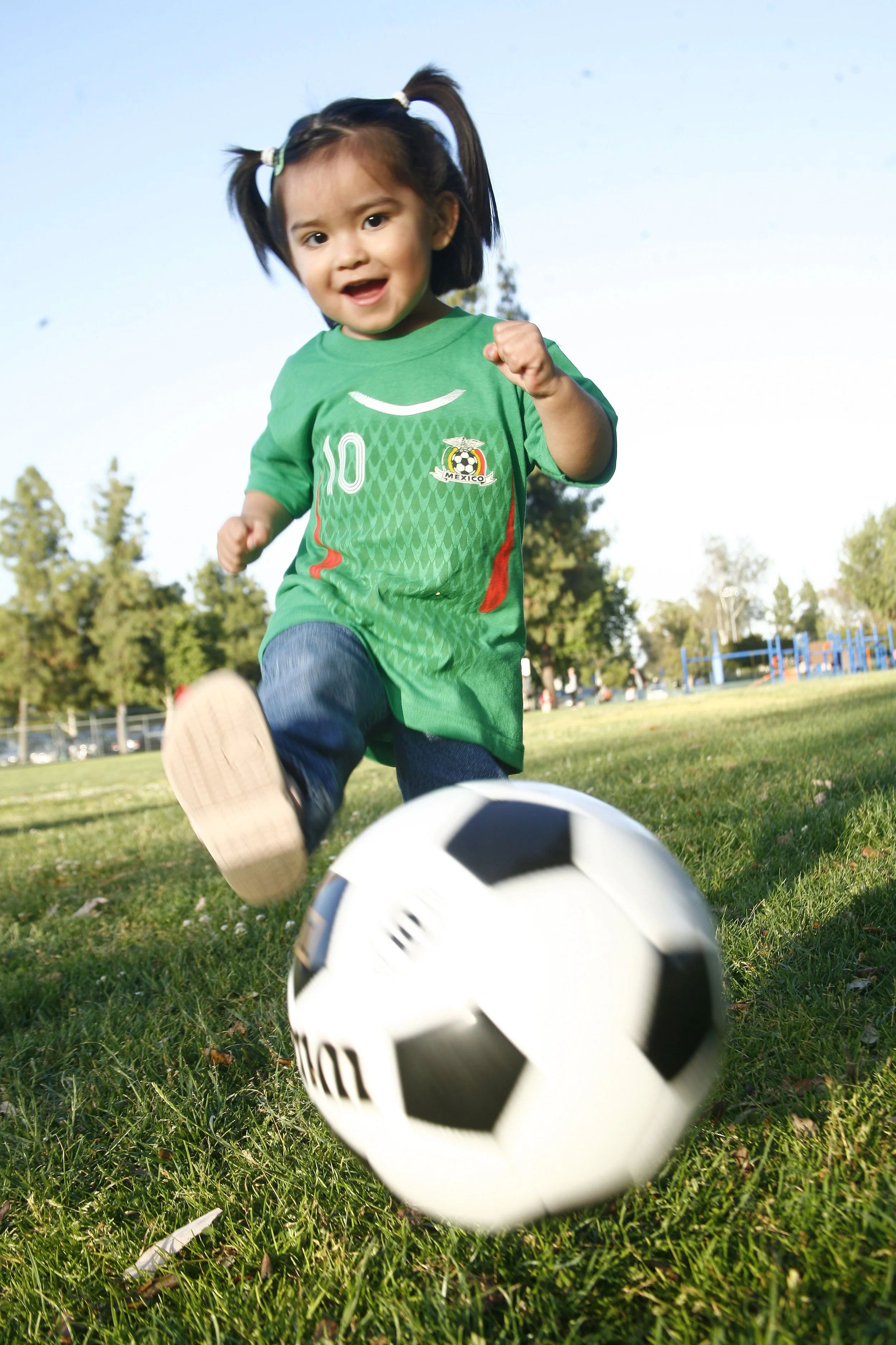 Young girl with pigtails wearing a Mexican soccer jersey kicking a soccer ball on a grassy field.