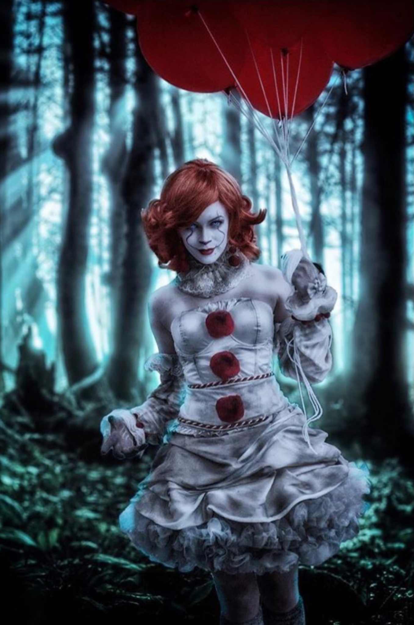 A person dressed as Pennywise from It in a forest with tall trees, holding red balloons, wearing a white ruffled dress with red pom-poms and clown makeup.