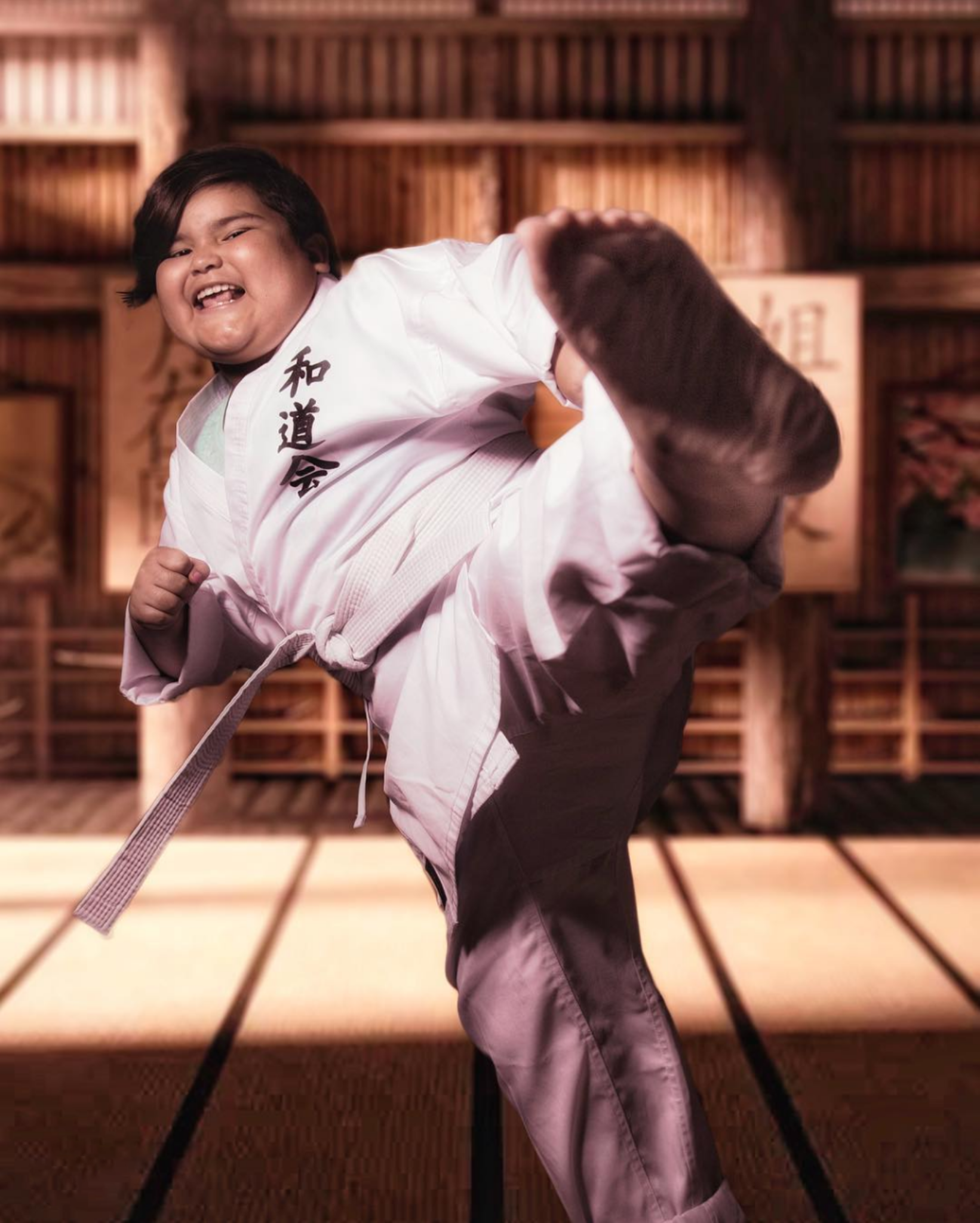 A young person with dark hair performing a martial arts move, wearing a white martial arts uniform with Japanese characters, in a dojo with wooden walls and tatami mats.
