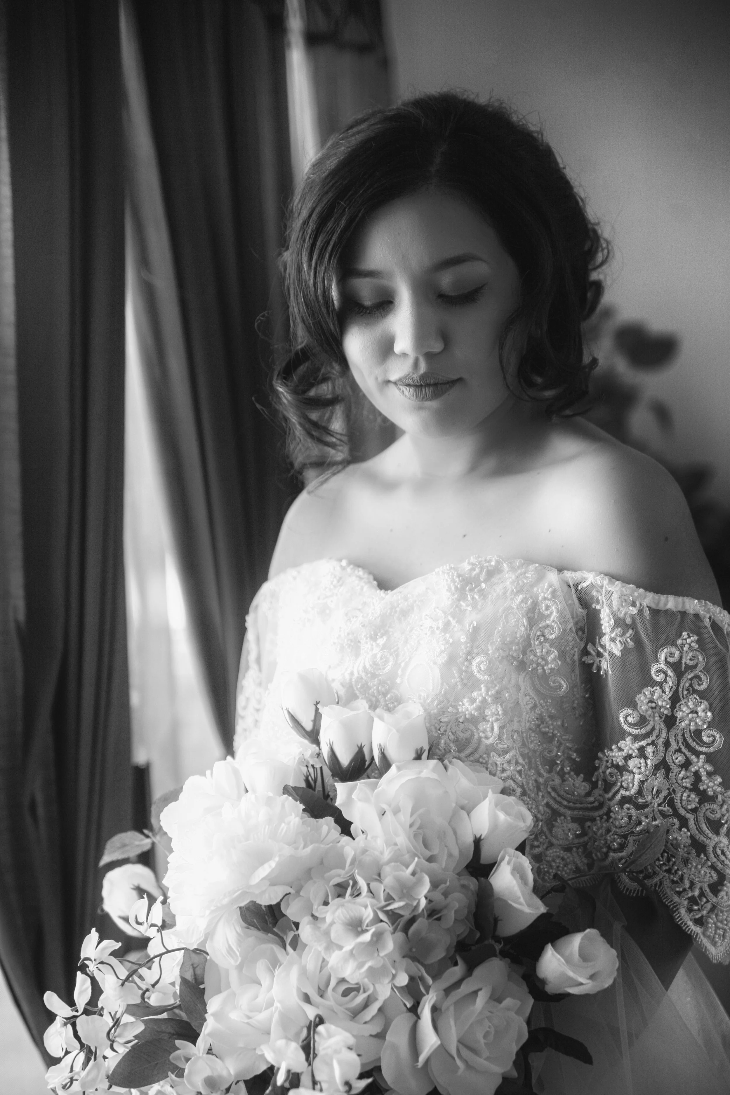 A black and white photograph of a woman in a wedding dress holding a bouquet of flowers, with curtains in the background.