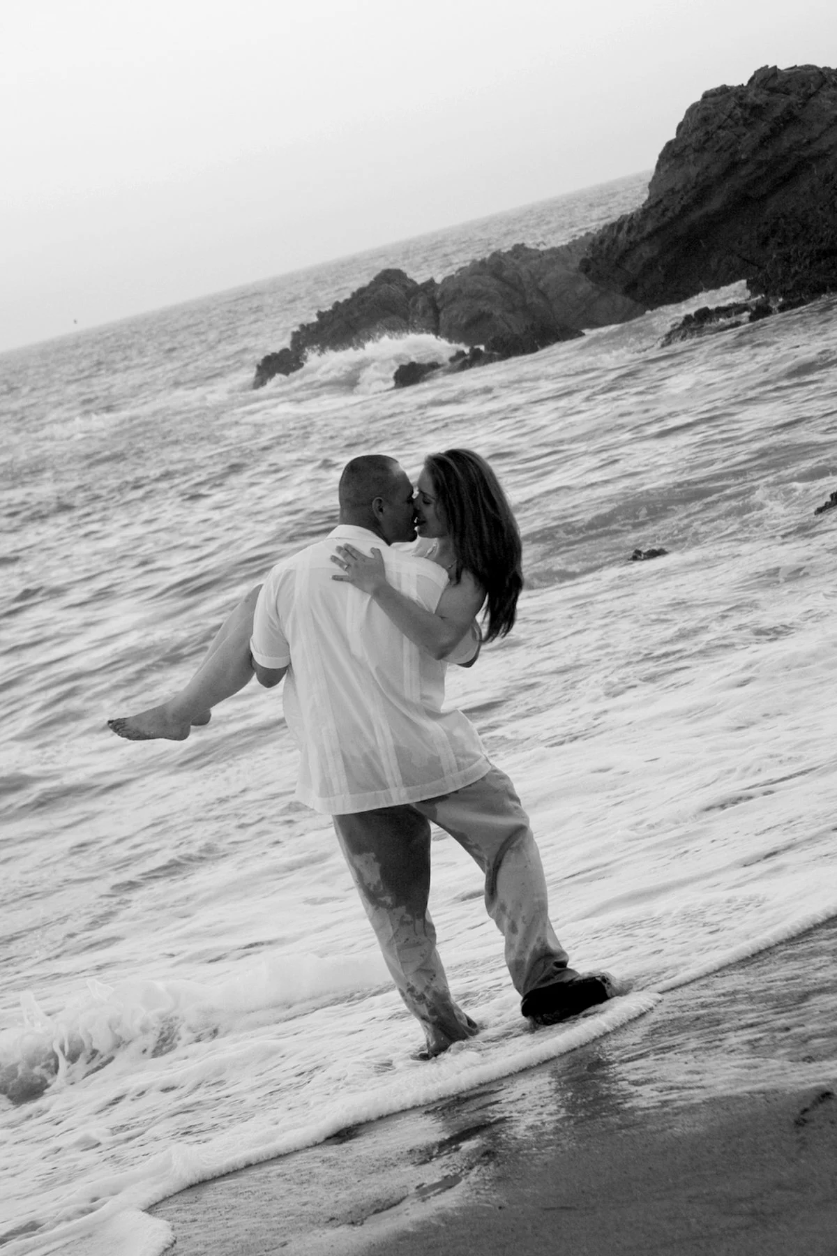 A couple sharing a romantic moment on the beach, with the man lifting the woman in his arms as they kiss, ocean waves and rocky cliffs in the background.
