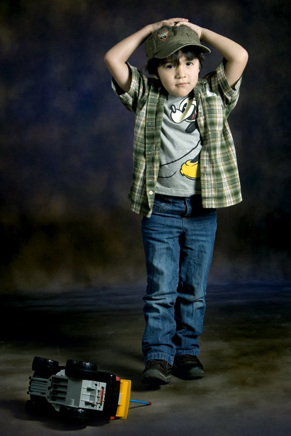 A young boy standing with his hands on his head, wearing a baseball cap, plaid shirt, graphic t-shirt, jeans, and sneakers, with a toy truck on the floor beside him, against a dark, studio background.