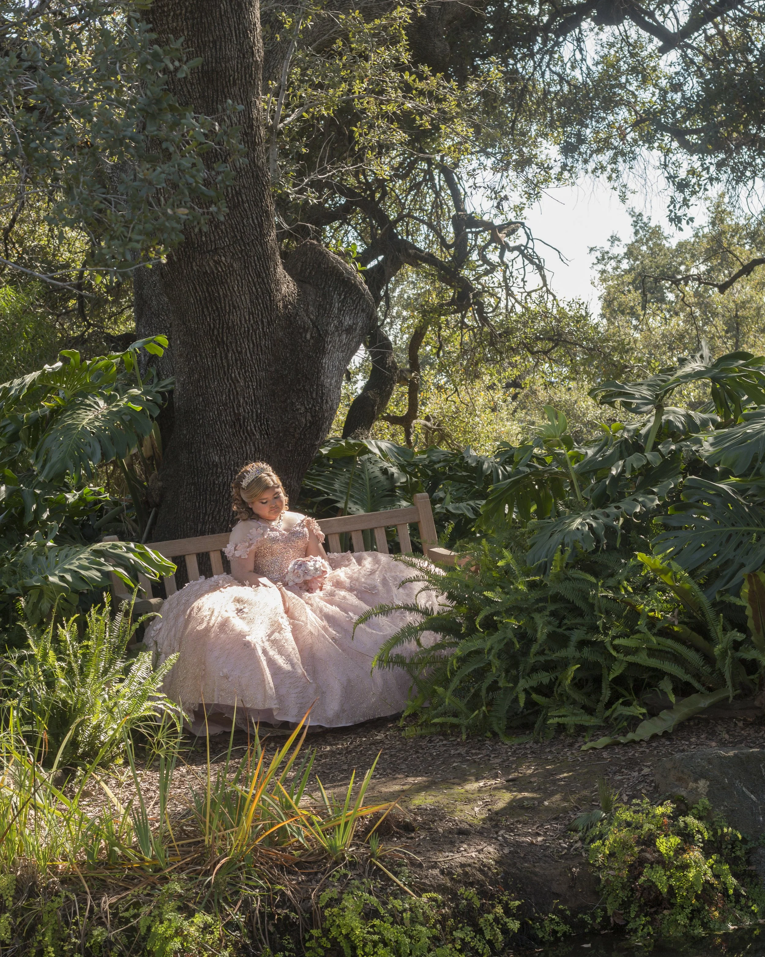 A young girl dressed in a formal gown and veil sitting on a bench outdoors in a forested area surrounded by lush green foliage and trees.