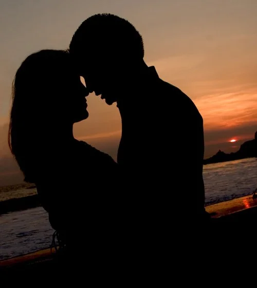 Silhouetted couple standing close together on beach at sunset.
