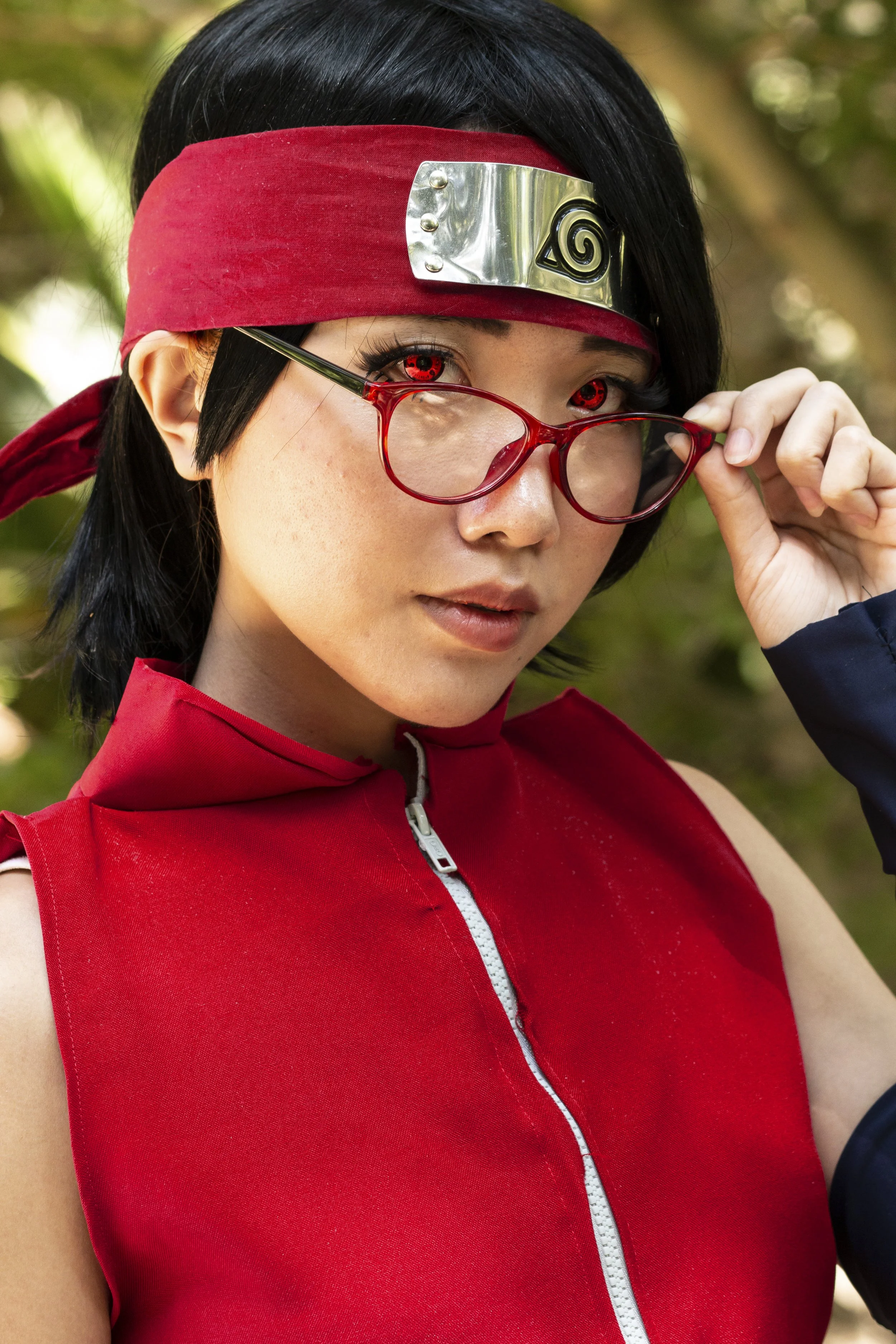 A woman with black hair, red contact lenses, and red glasses, wearing a red headband with a silver metallic forehead protector featuring a black spiral symbol, and a red sleeveless vest with a white zipper, posing outdoors.