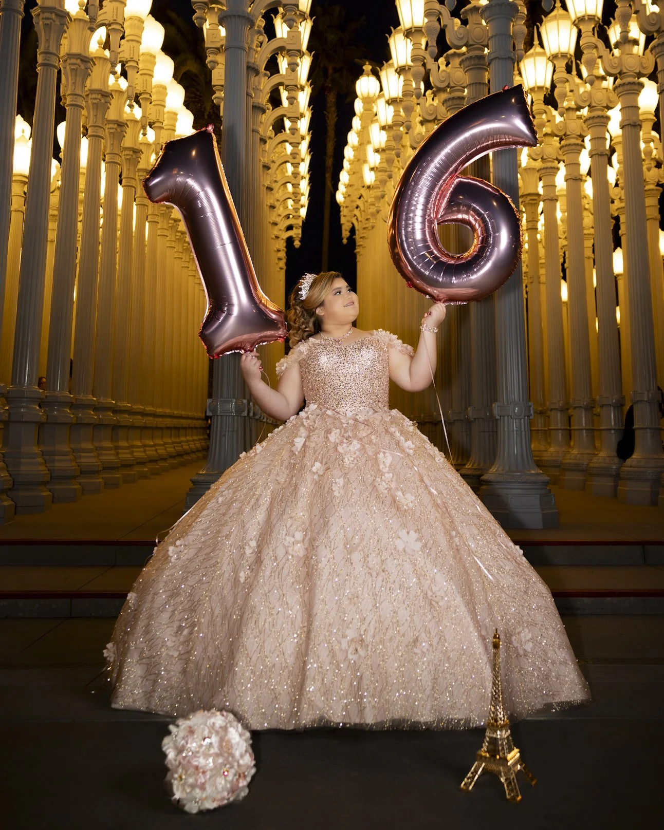 A young girl in a peach-colored ball gown holding large pink balloons that form the number six, standing in front of a decorative wall of illuminated lamp posts at night.