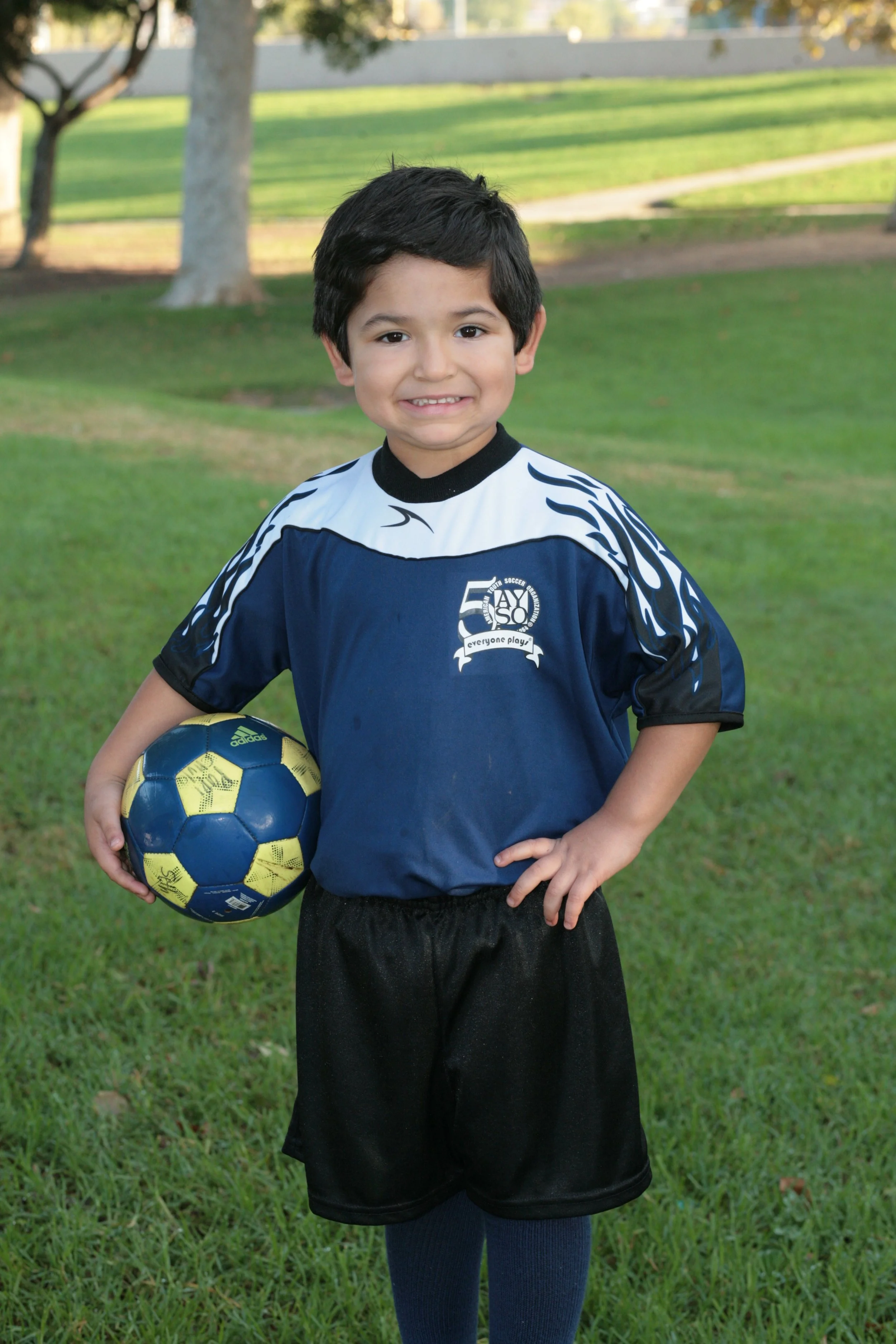 Young boy wearing a sports jersey and shorts holding a blue and yellow Adidas soccer ball, standing on a grassy park with trees in the background.