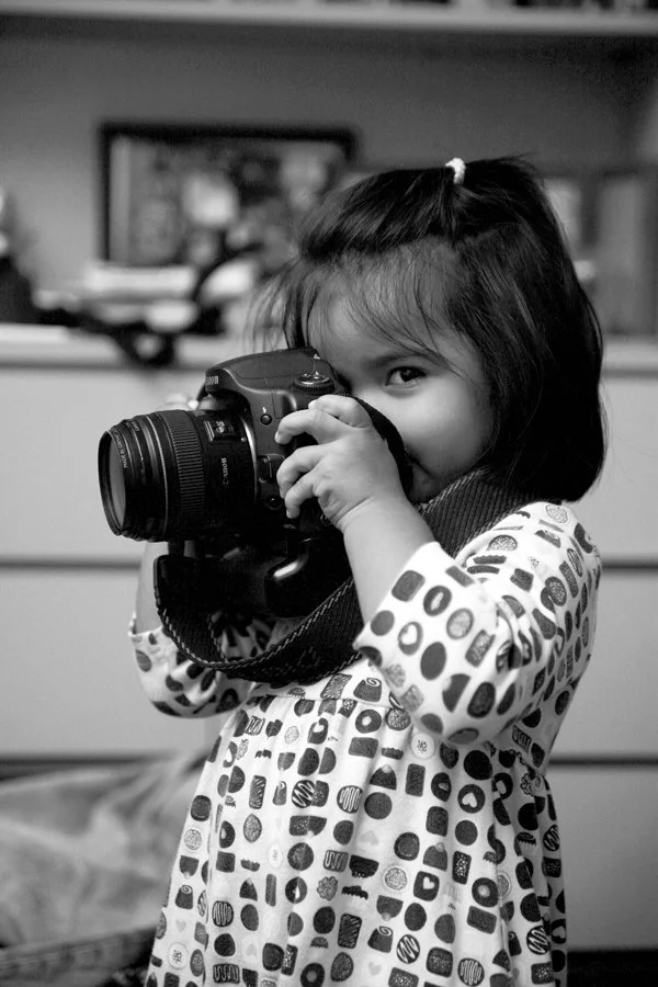A young girl with shoulder-length hair, wearing a polka dot dress, holding a camera to her eye as she looks through the viewfinder.
