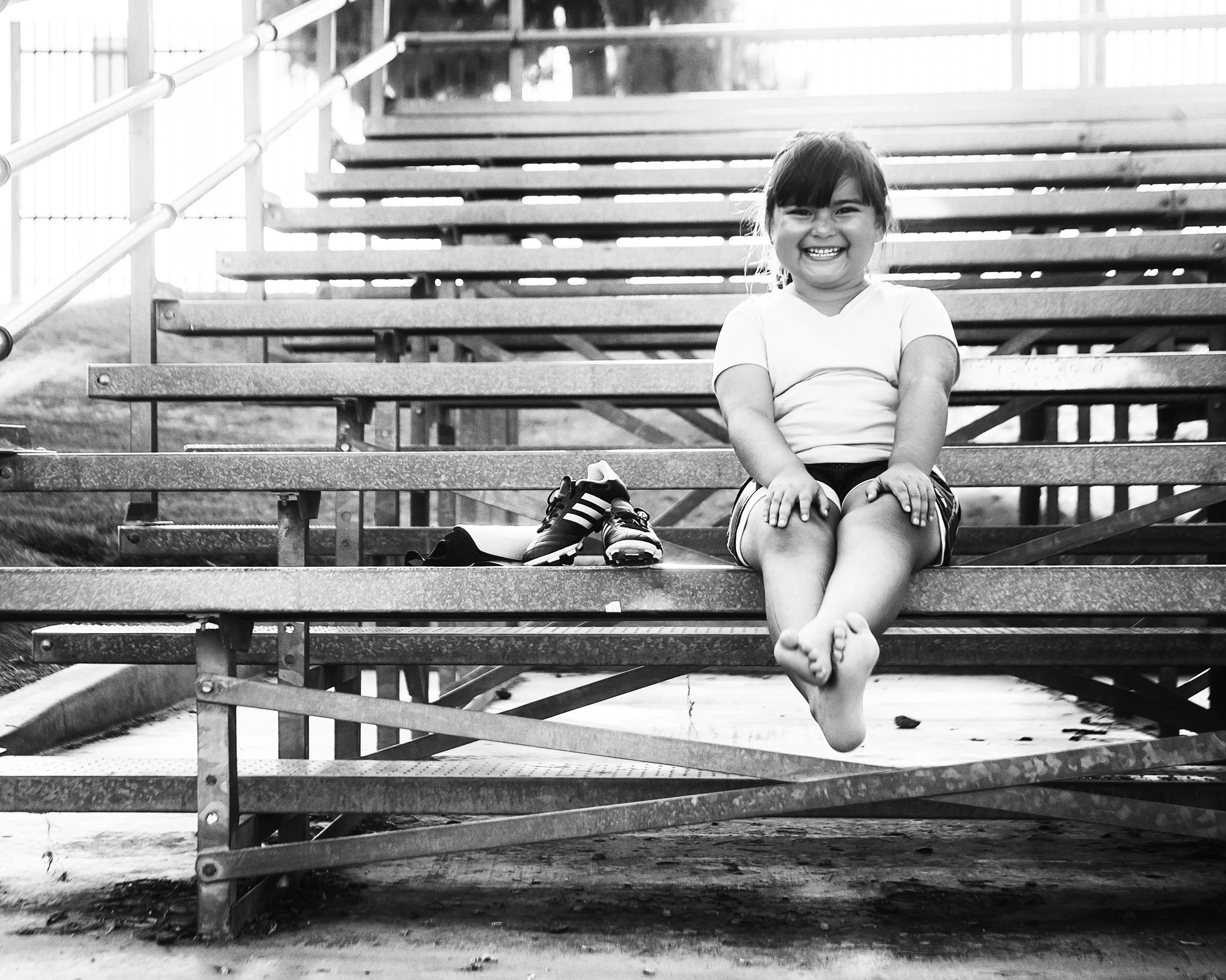 A young girl sitting on metal bleachers, smiling and showing her teeth, with her shoes placed on the step in front of her. She is wearing a light-colored shirt and shorts, and has dark hair with bangs. The background includes more bleacher steps and a railing.