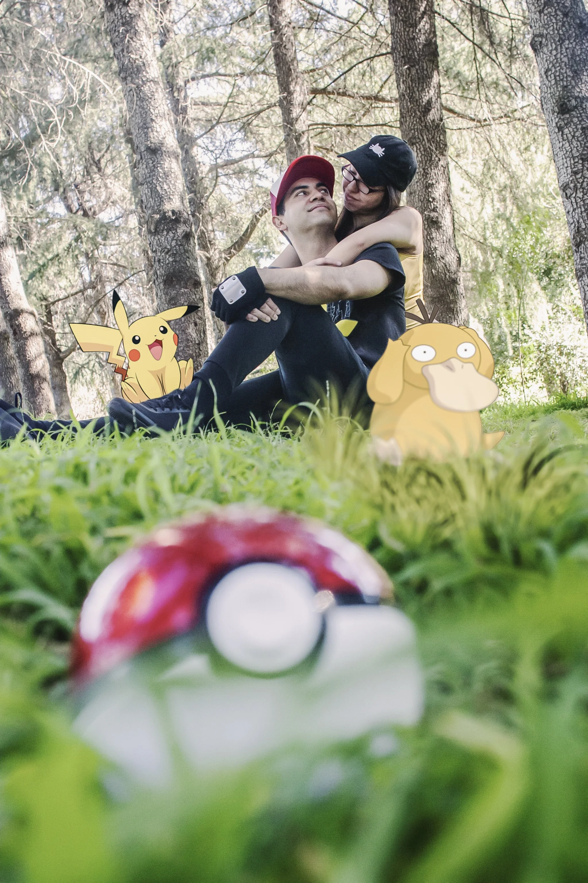 A photograph of a young man and woman sitting on the grass in a wooded area, with cartoon Pokémon characters Pikachu and Dugtrio placed in the foreground. The young man is wearing a black shirt and a red cap, and the young woman is wearing glasses and a black hat. They are hugging and looking at each other.