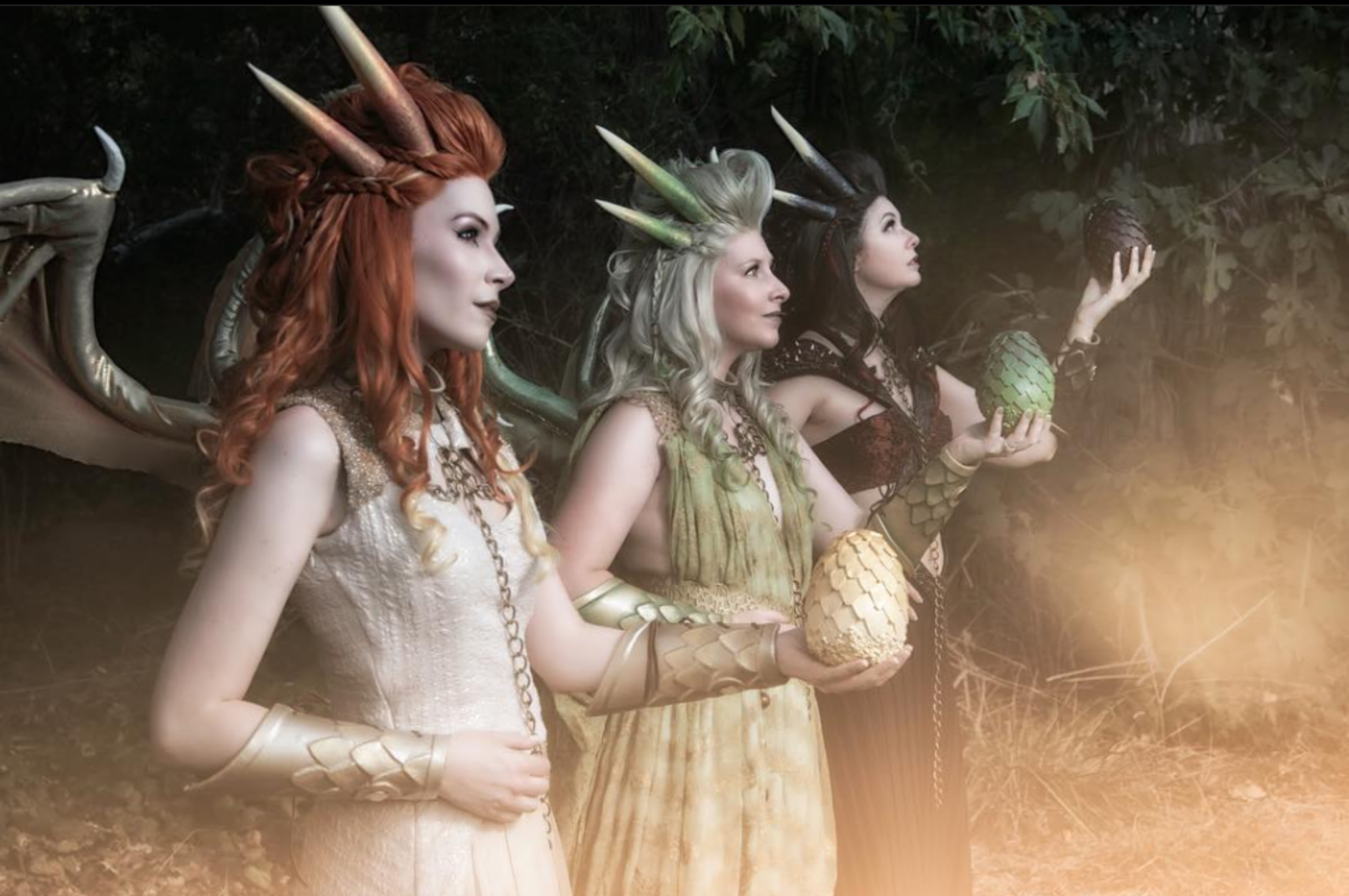 Three women dressed as fantasy characters with unicorn horns and scales on their arms, holding pineapples, in a forest setting.