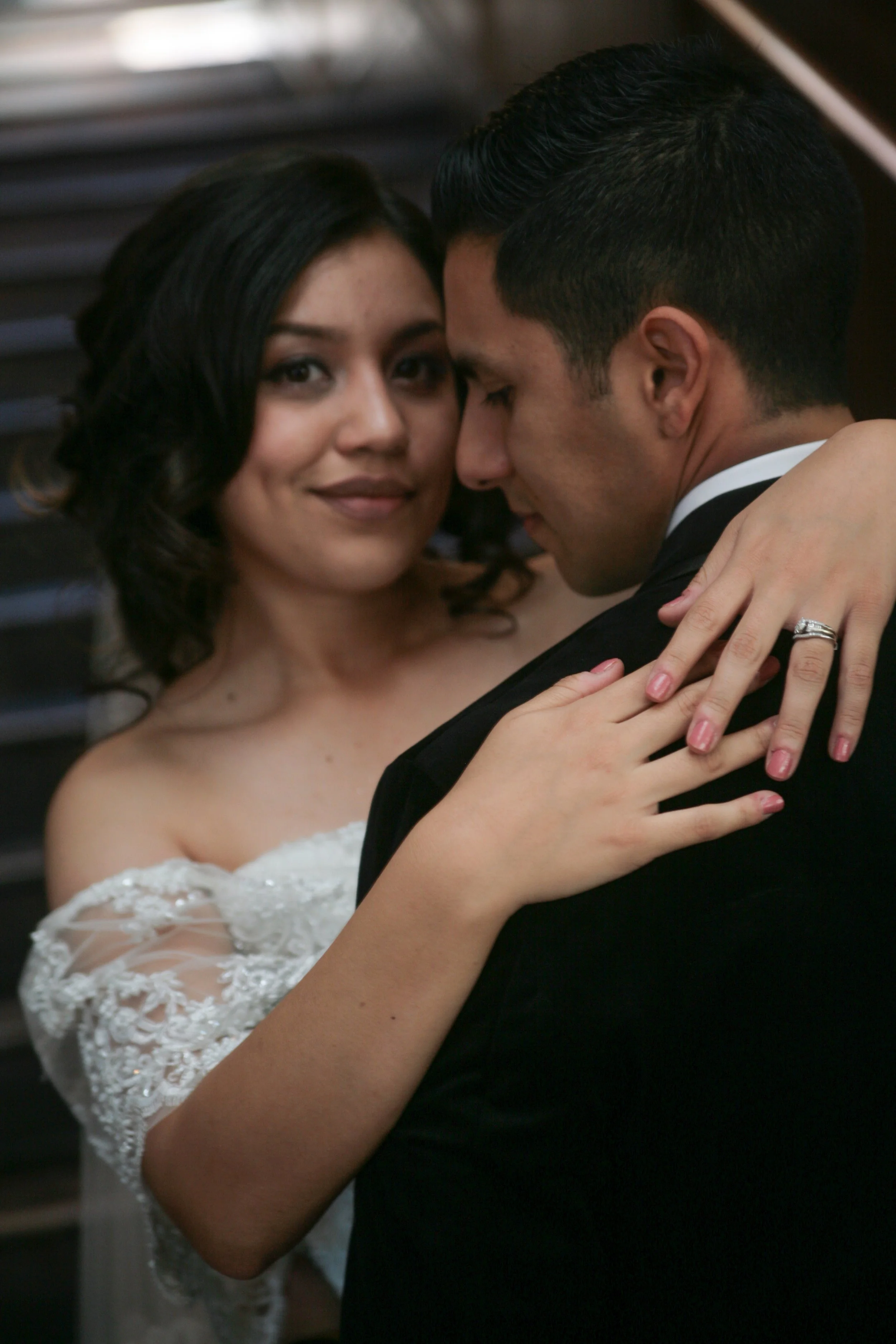 A bride and groom are dancing closely, with the bride smiling at the camera and the groom leaning in with his eyes closed. The bride is wearing a white lace wedding dress and the groom is in a black tuxedo.