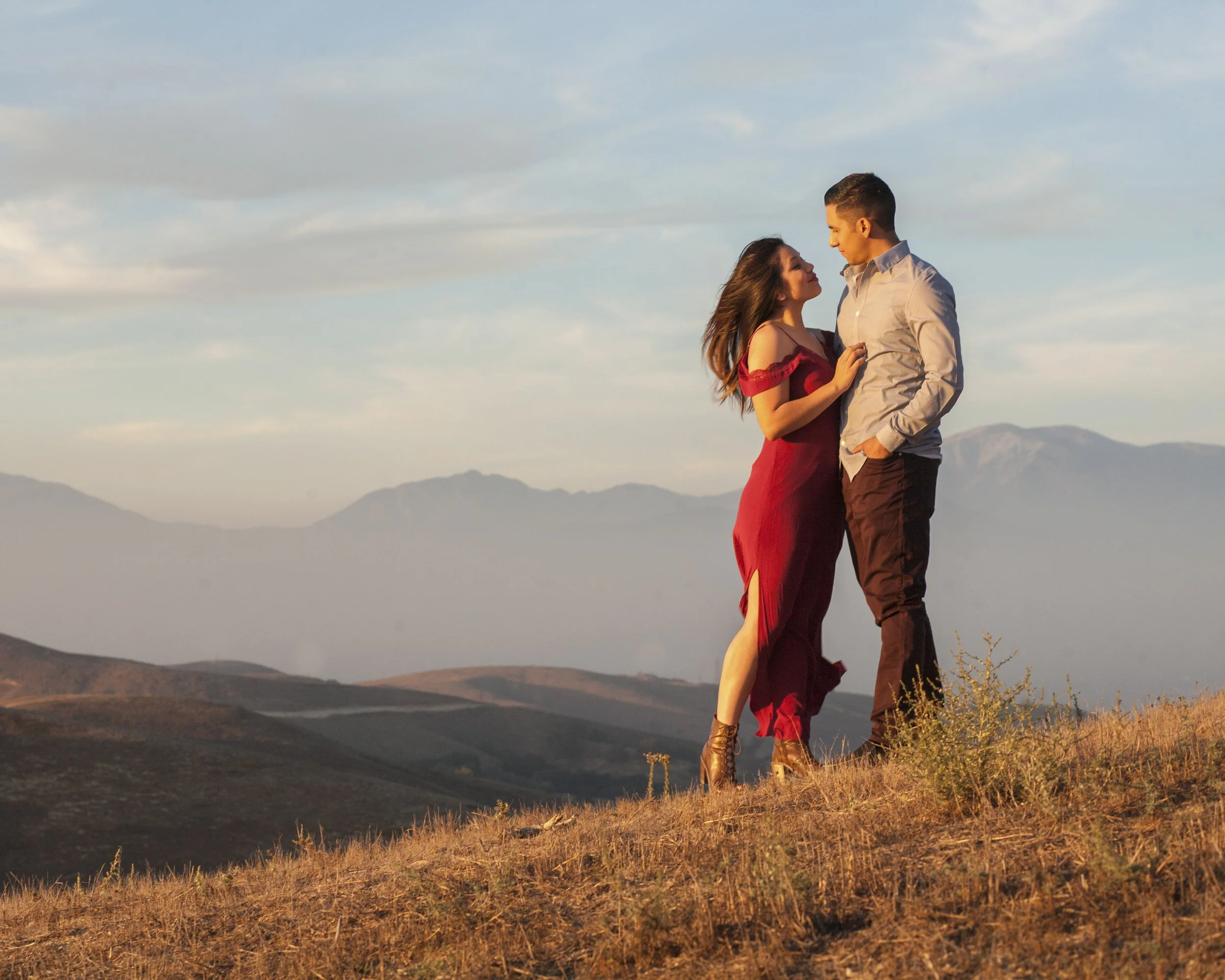 A young couple stands close together on a hilltop during sunset. The woman, wearing a long red dress with shoulder cut-outs and ankle boots, gazes into the man's eyes. The man, dressed in a button-up shirt and dark pants, looks back at her. They are surrounded by dry grass with mountain ranges and a partly cloudy sky in the background.