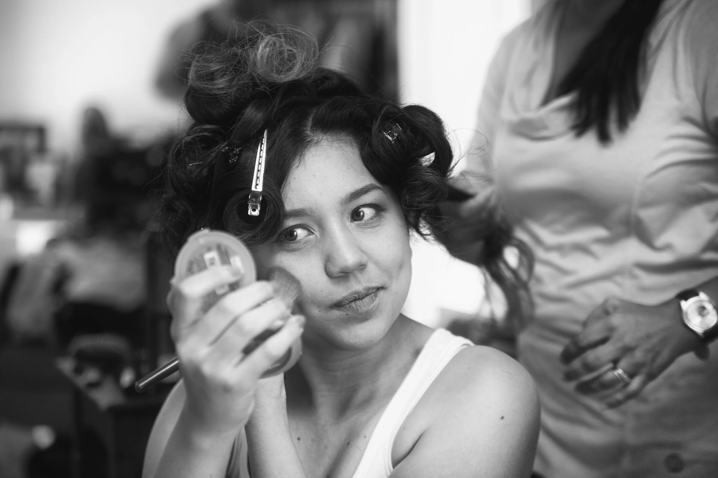 A woman with dark curly hair is having her hair styled with rollers and clips by a hairstylist. She is looking towards the camera while the stylist works on her hair.