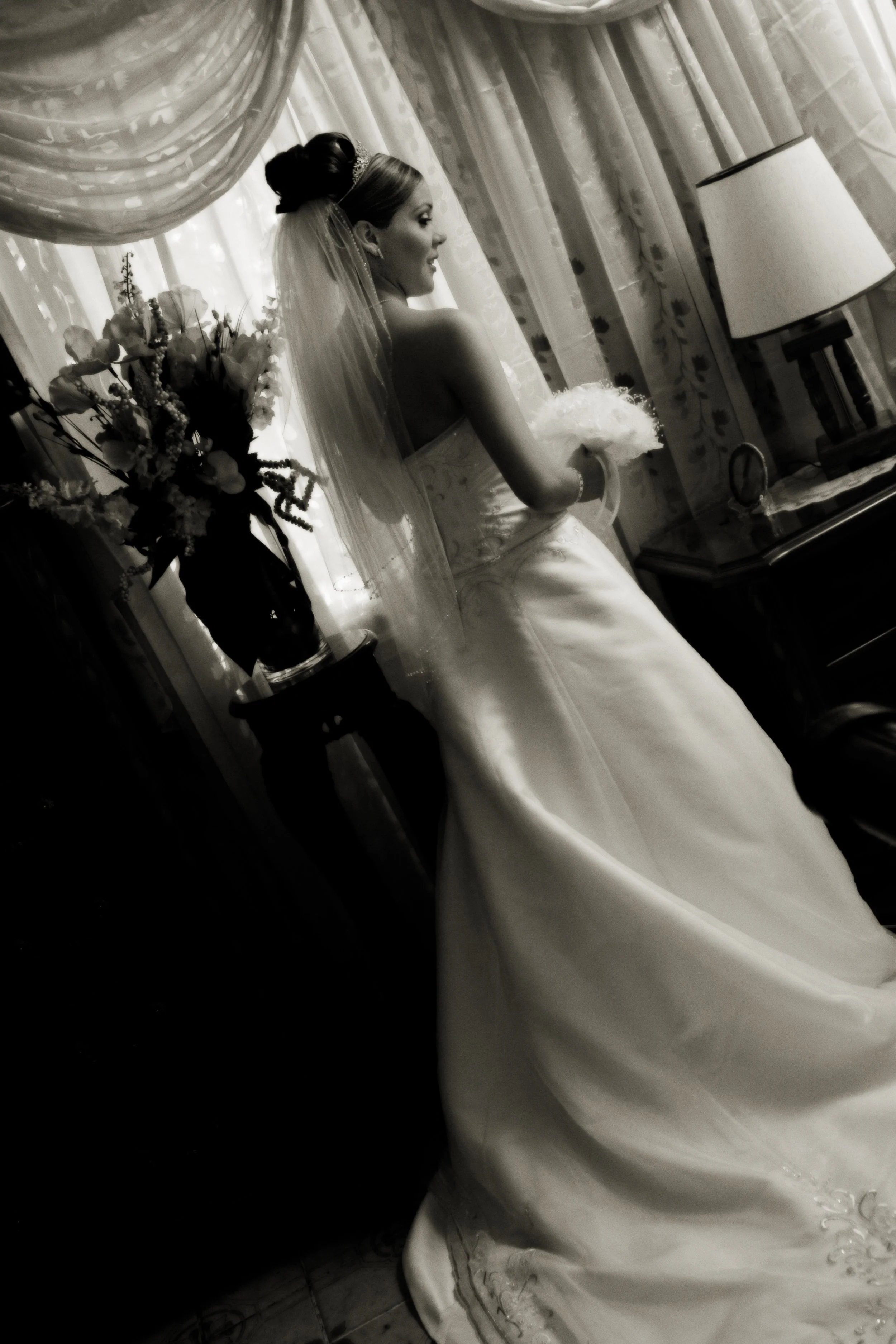 A bride in a wedding gown with a veil, holding a bouquet, standing indoors near a window with floral curtains, next to a table with a lamp and flowers.