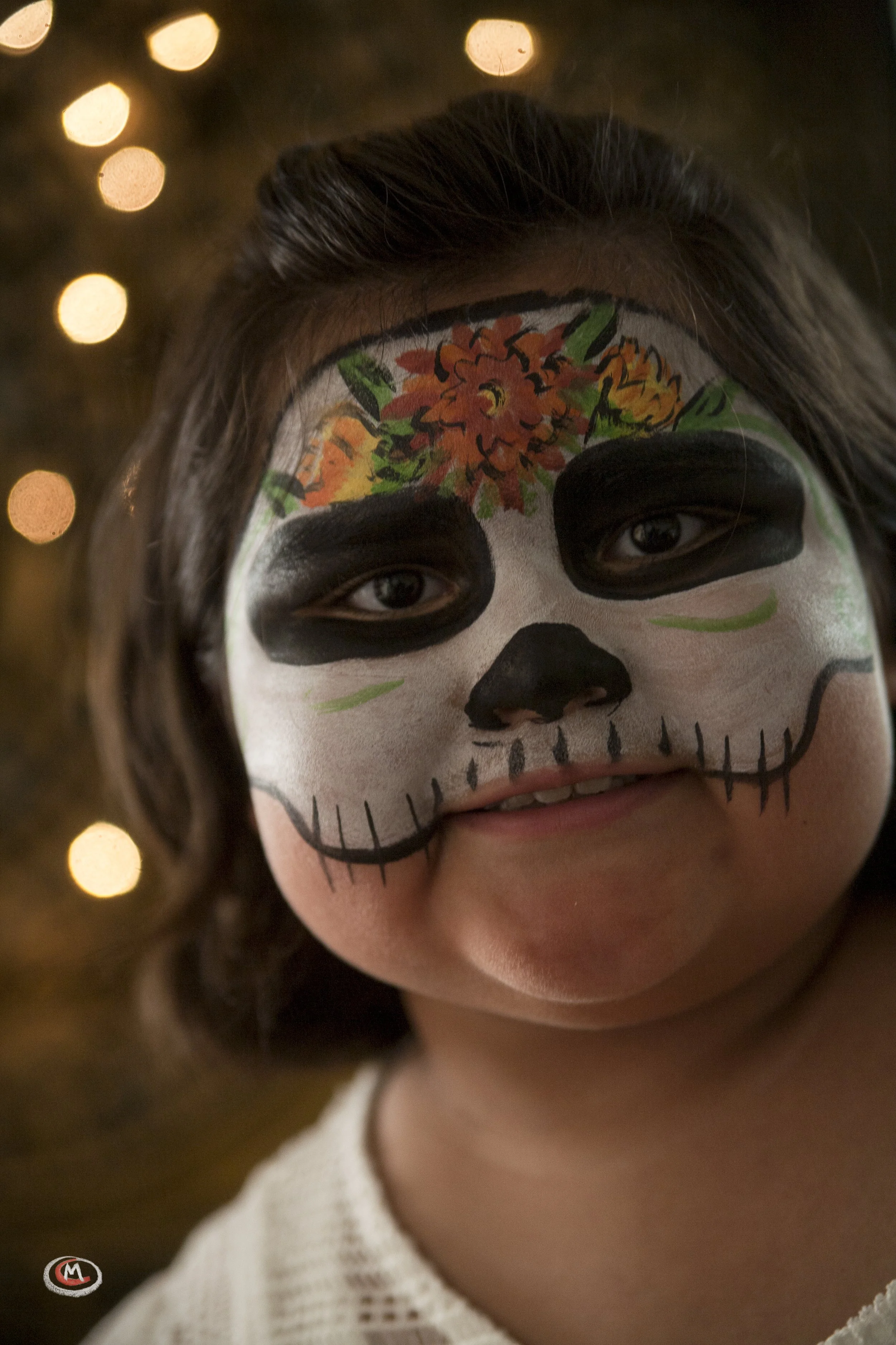 A young girl with face paint resembling a sugar skull, with orange flowers and green leaves on her forehead, black eye sockets, and a black nose. She is smiling slightly and has dark hair. The background features blurred warm lights.