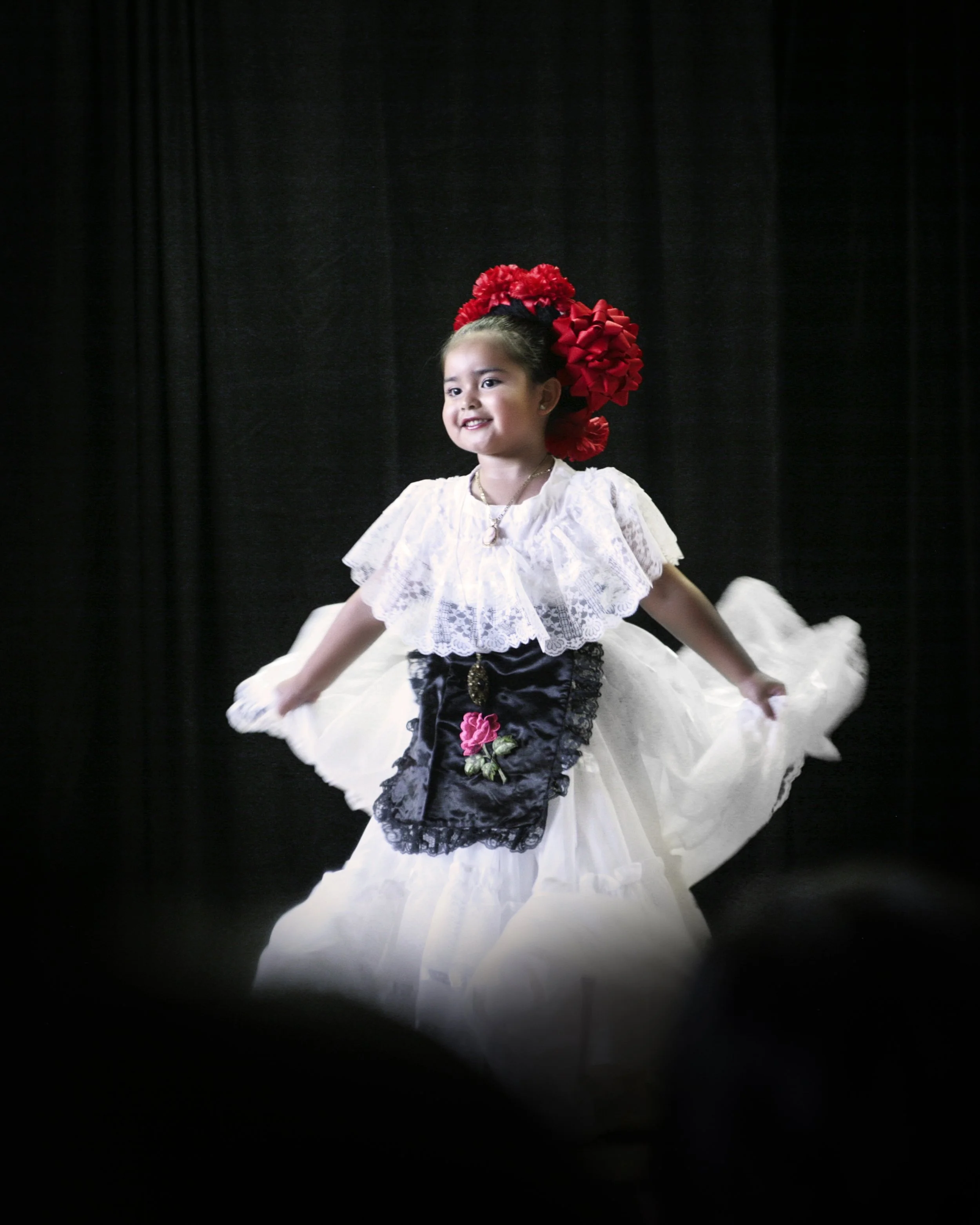 Young girl in traditional Spanish dress dancing on stage with black curtain background.