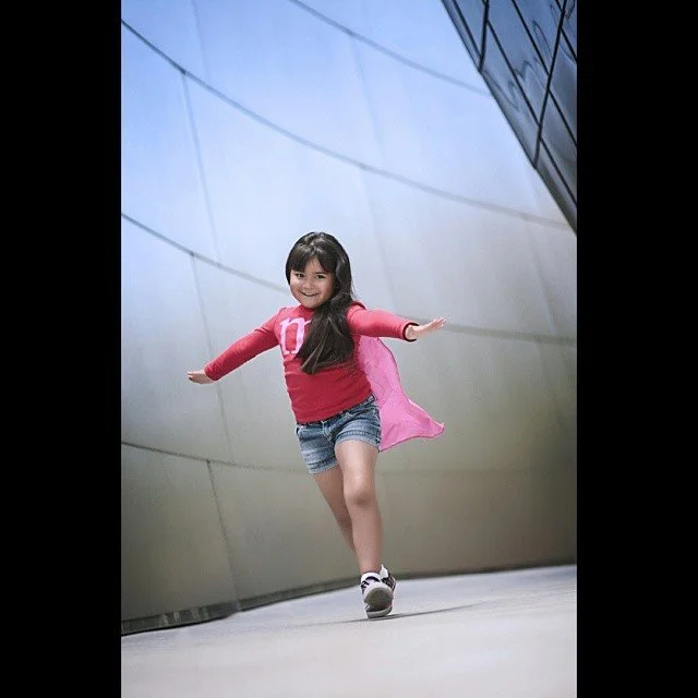 A young girl with long dark hair, smiling and wearing a pink jacket, red shirt, denim shorts, and white sneakers, walking with arms outstretched on a smooth, curved metallic surface at a modern building or structure.