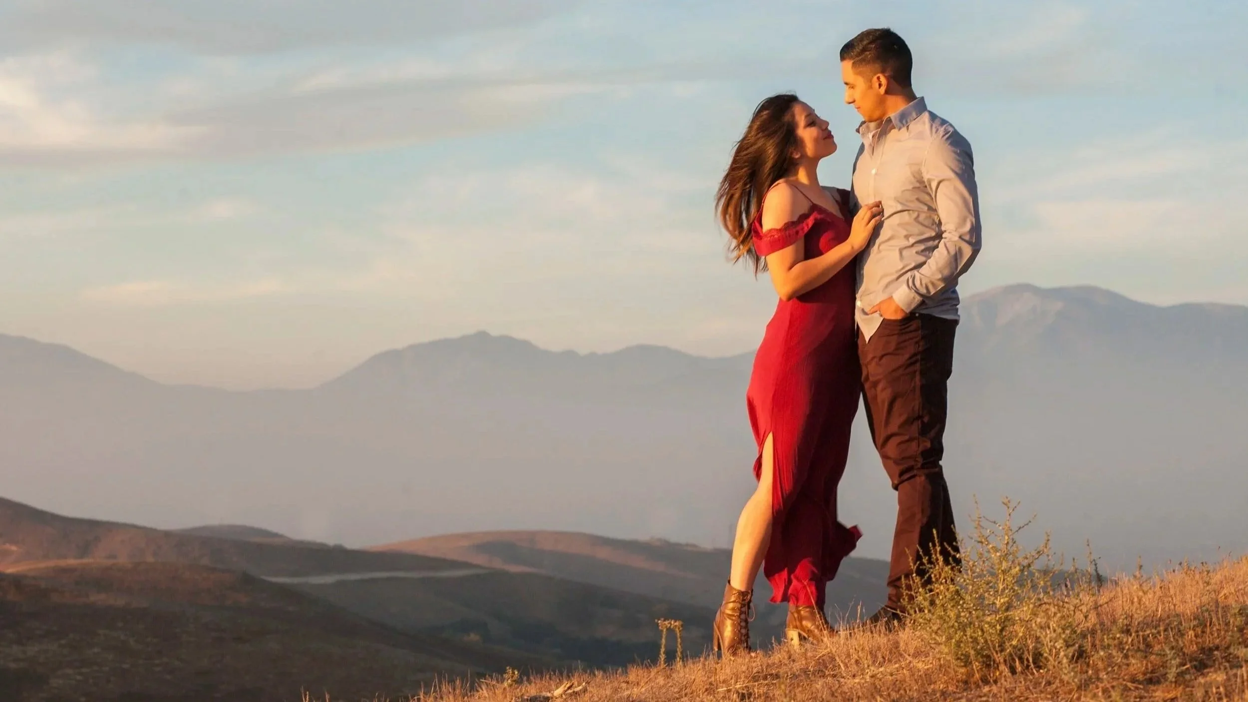 A couple stands on a hilltop during sunset, with mountains in the background. The woman is wearing a red dress and the man is in a light-colored shirt and dark pants. They are looking at each other romantically.