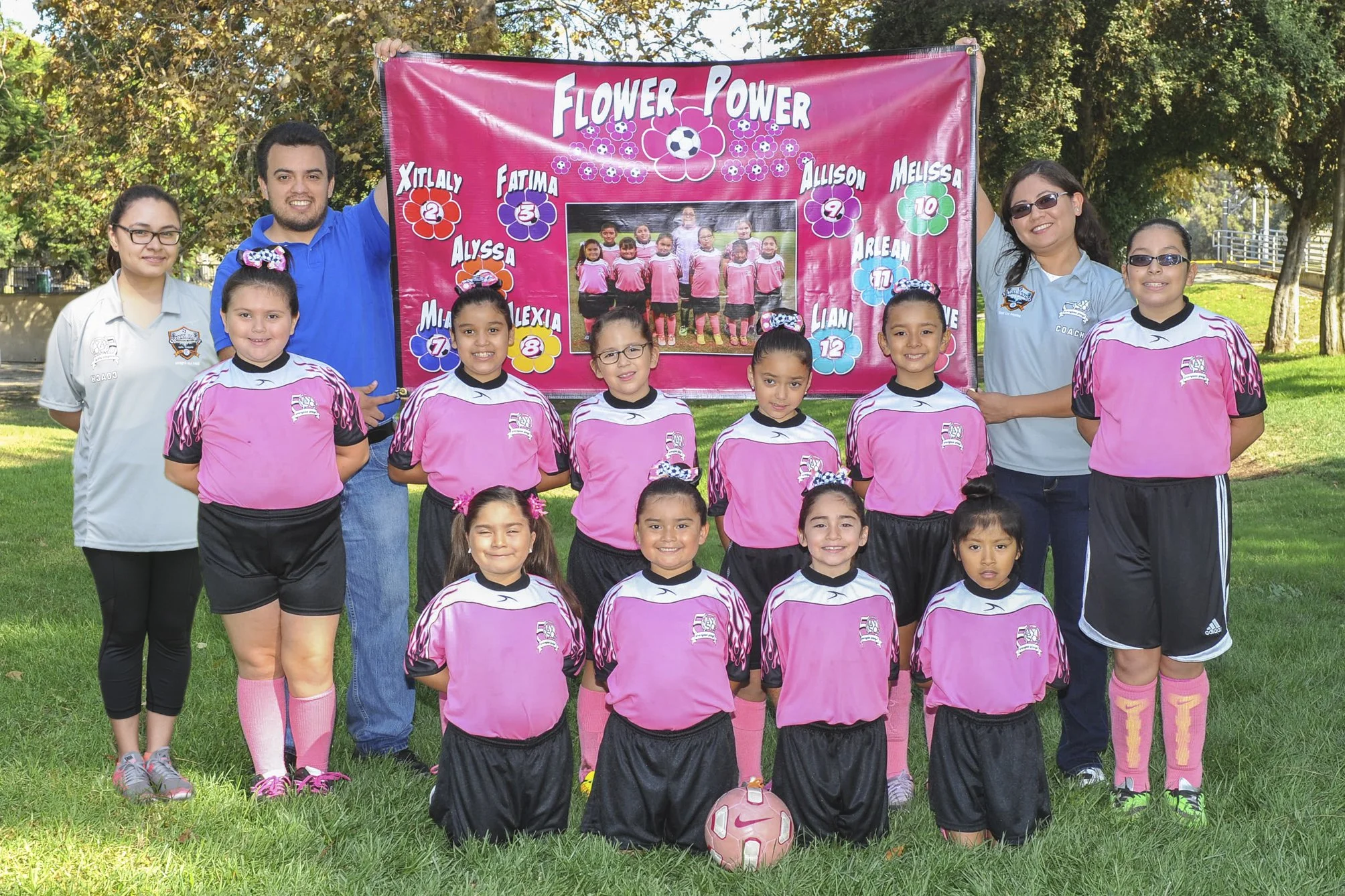 A youth soccer team and their coaches standing outdoors in front of a pink banner with flowers, a soccer ball, and team names. The team is wearing pink and black uniforms, and there is a soccer ball on the grass in front.