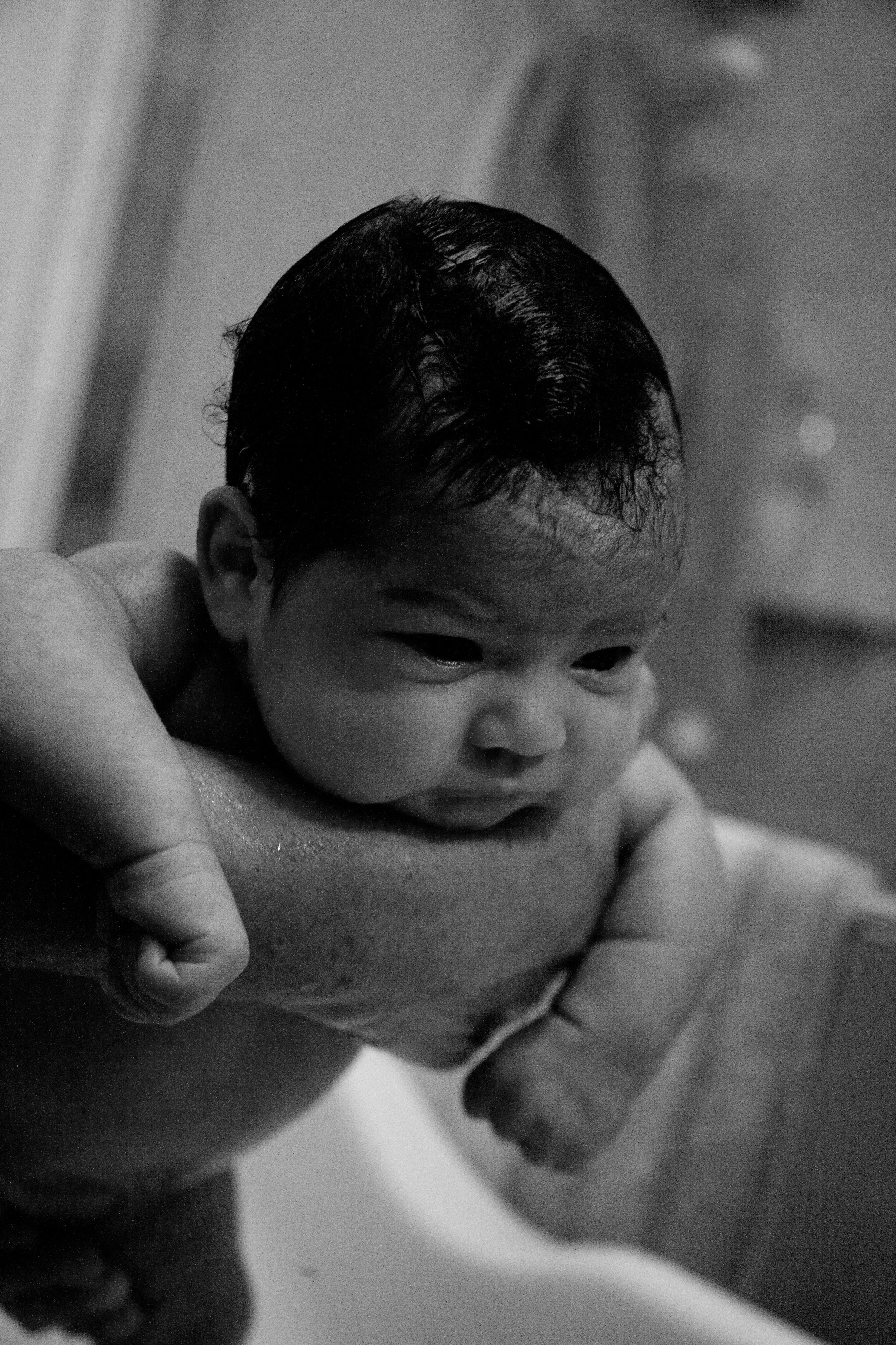 Close-up of a baby's face with dark hair, showing only the upper part of the face, held in an adult's hand in a black-and-white photo.