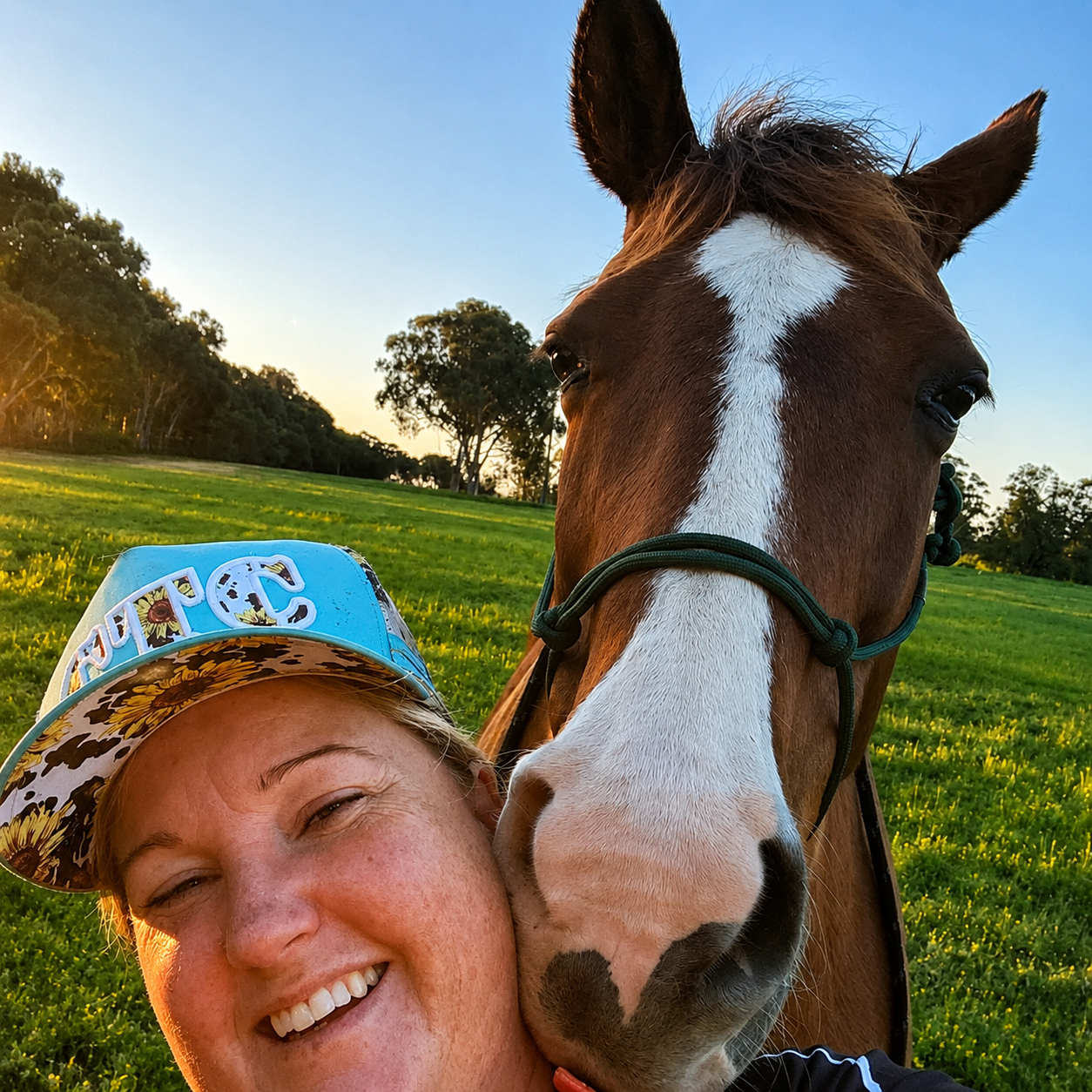 A woman with a big smile taking a selfie with a brown and white horse in a grassy field during sunset.