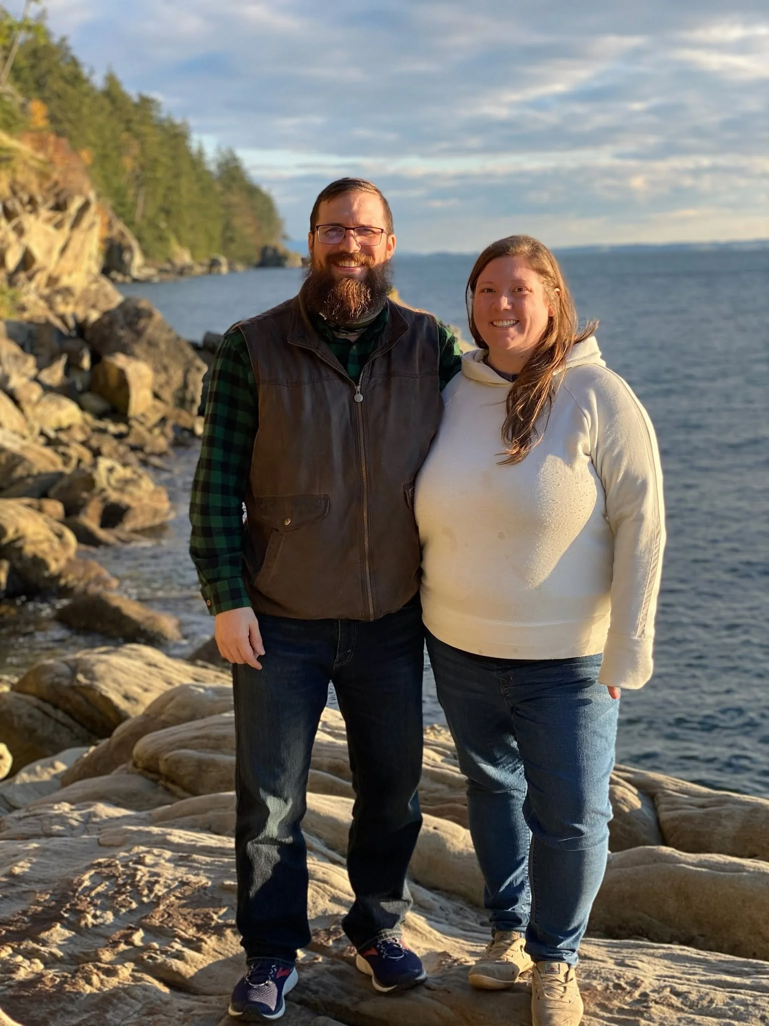 A man and woman standing on rocks near a body of water, smiling at the camera, with trees and a cloudy sky in the background.