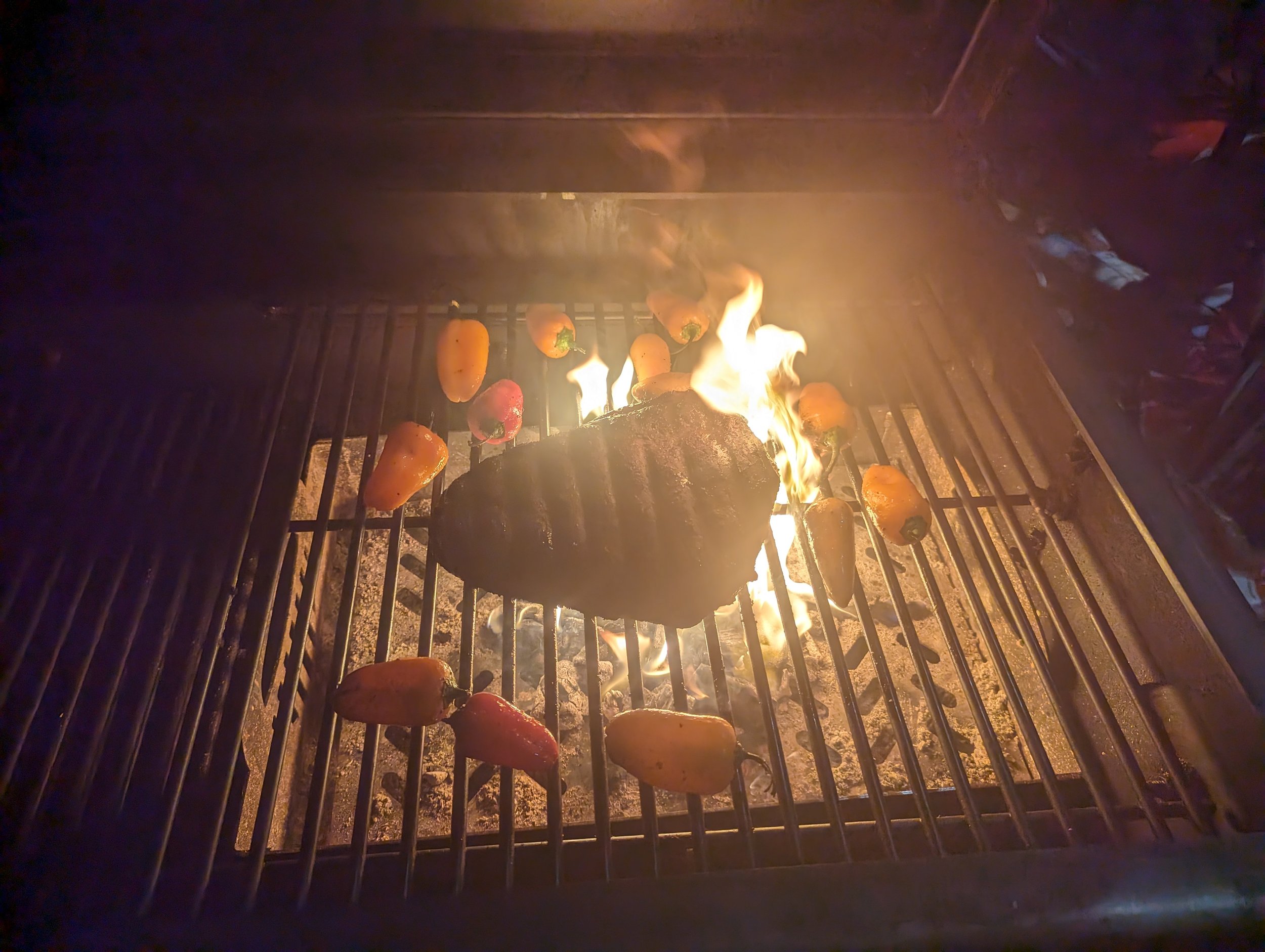 Meat and colorful peppers cooking on a grill with open flames at night.