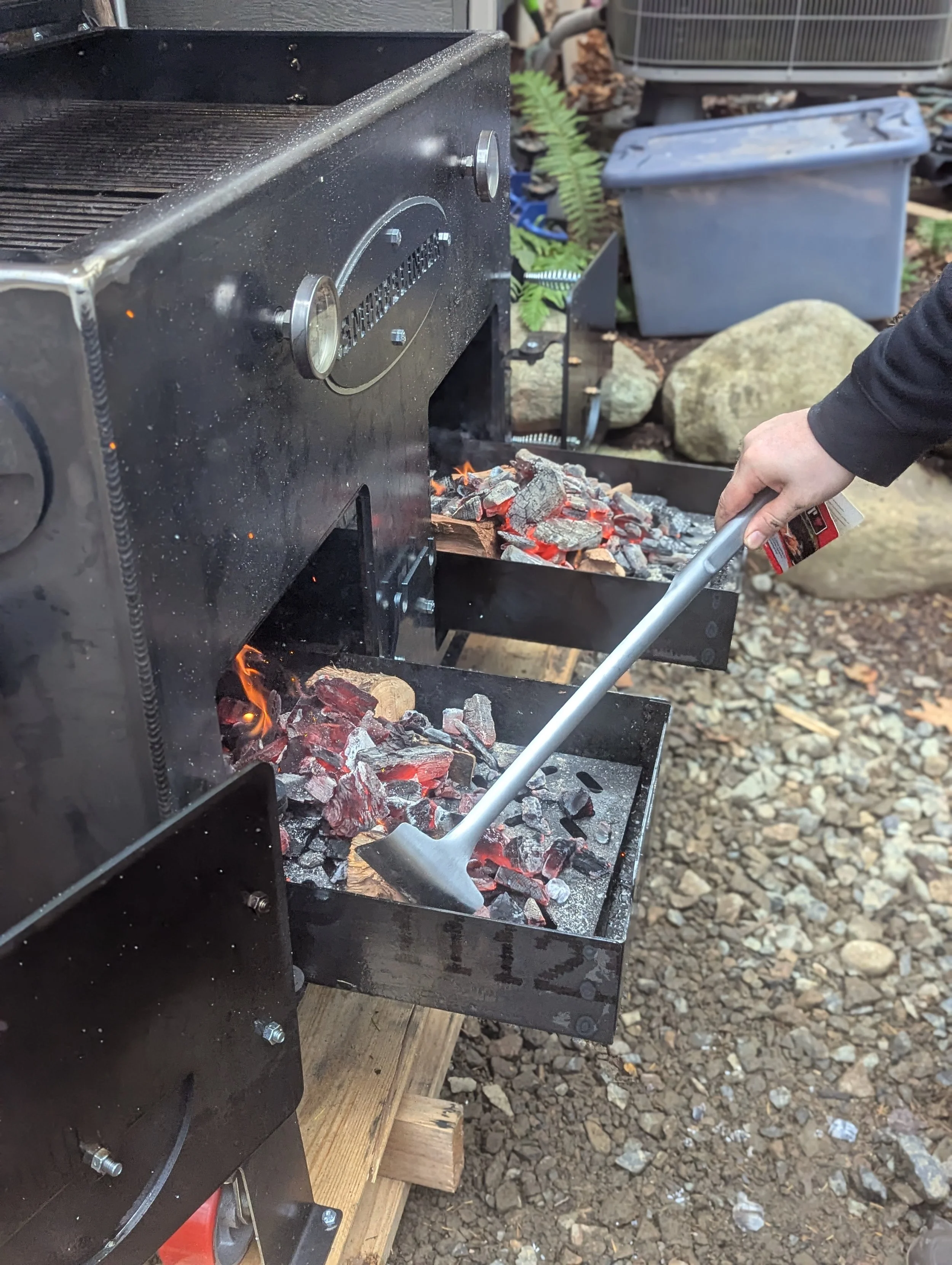 A charcoal grill with smoke rising from the coals, situated outdoors on a wooden platform, surrounded by rocks, potted plants, and outdoor storage containers.