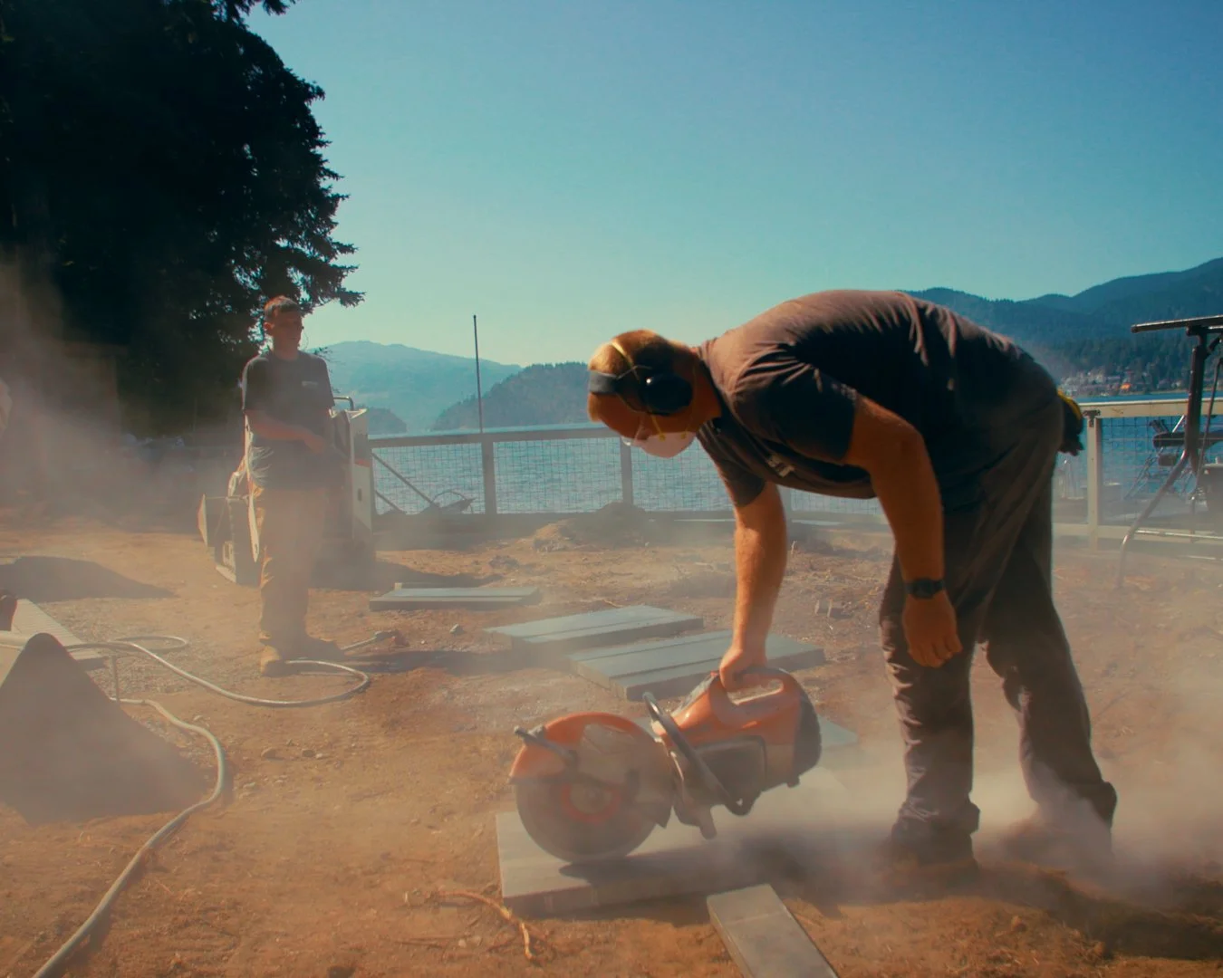 A man using a chainsaw on a wooden plank outdoors near a body of water, with mountains in the background and another person standing nearby.