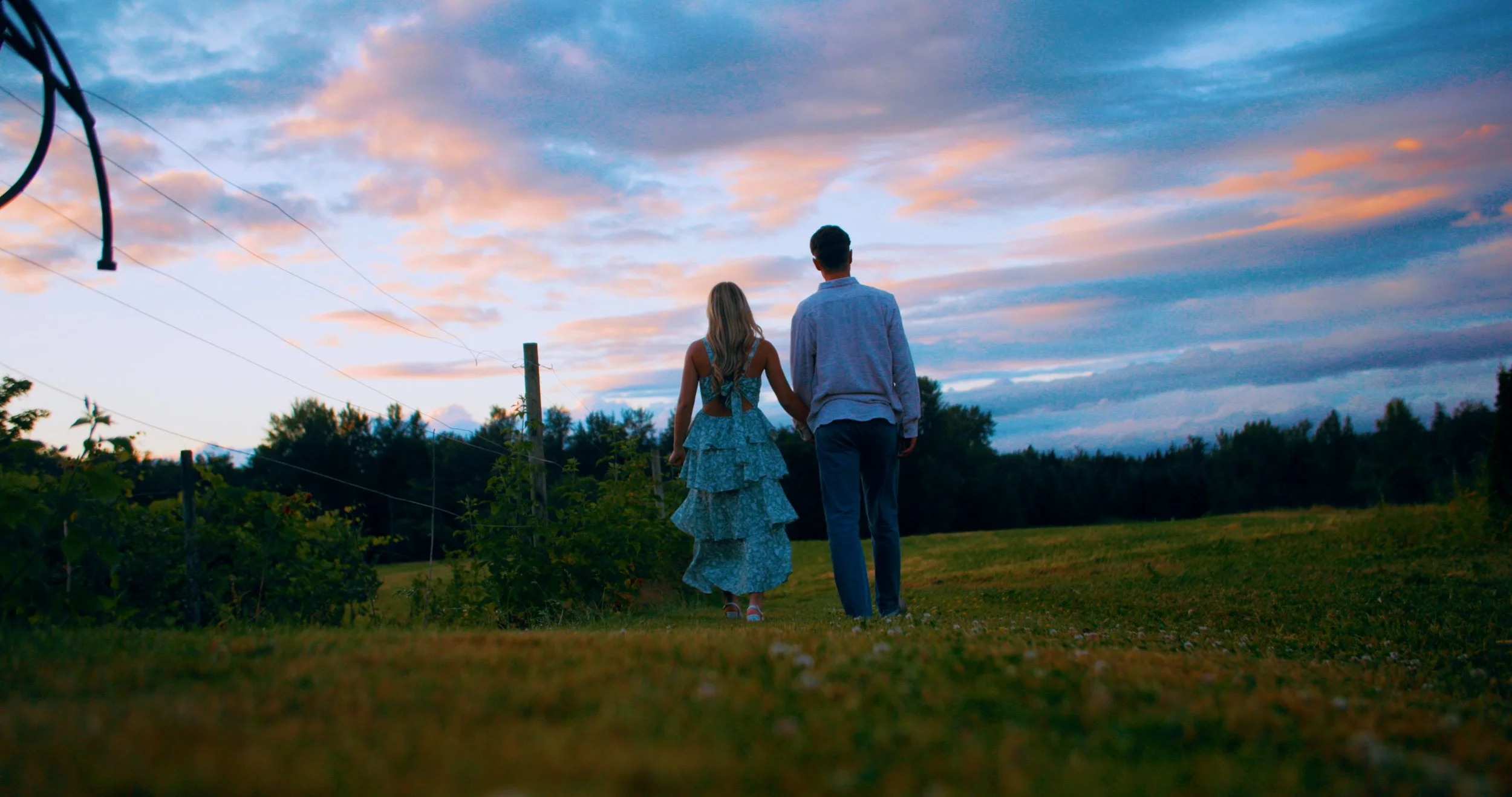 A couple holding hands walking in a field during sunset, with colorful clouds in the sky and trees in the background.