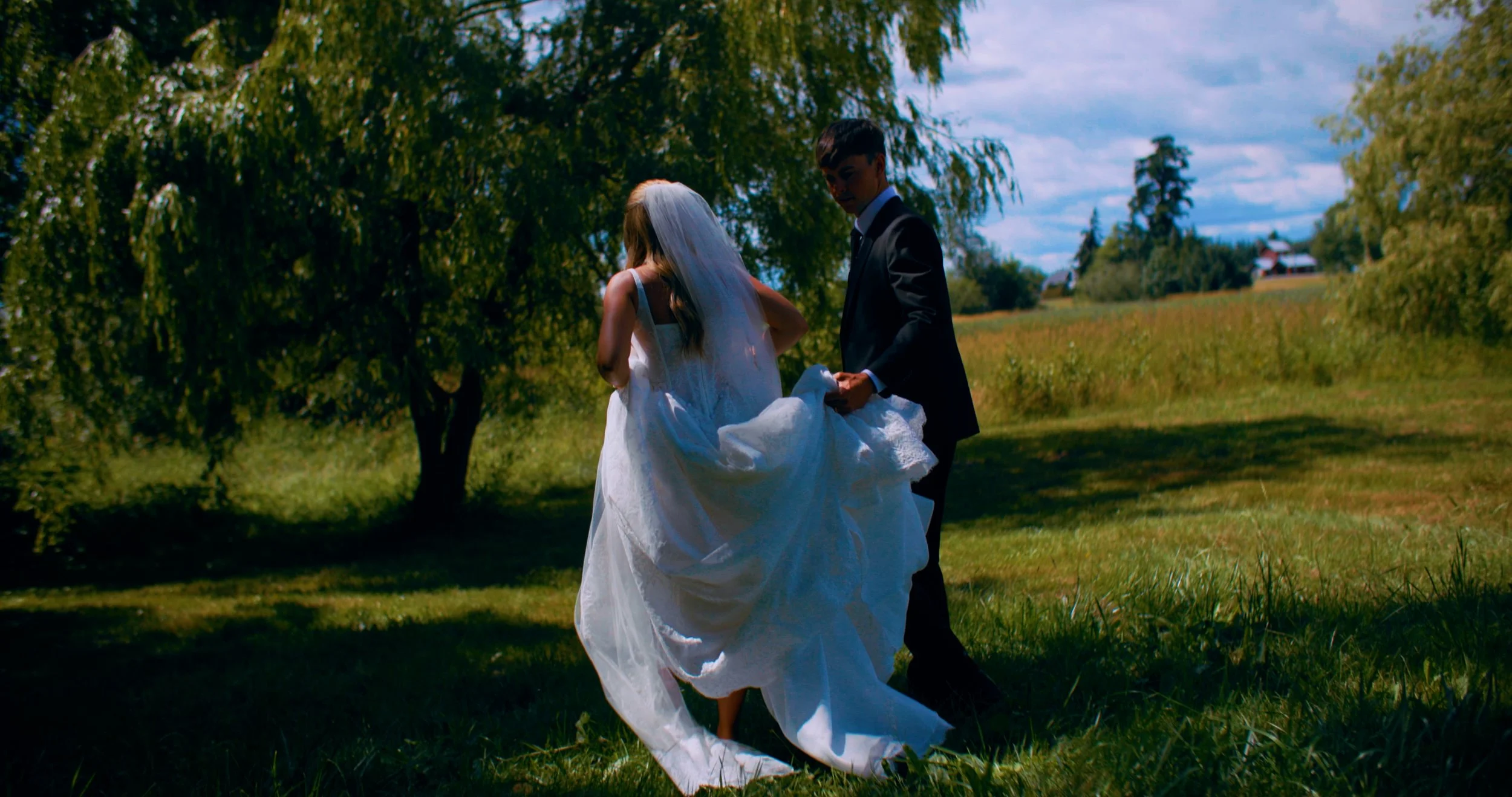 A bride in a white wedding dress and veil holding a swan-shaped pillow, walking on grass with a groom in a black suit beside her in a park-like setting with trees and a field.