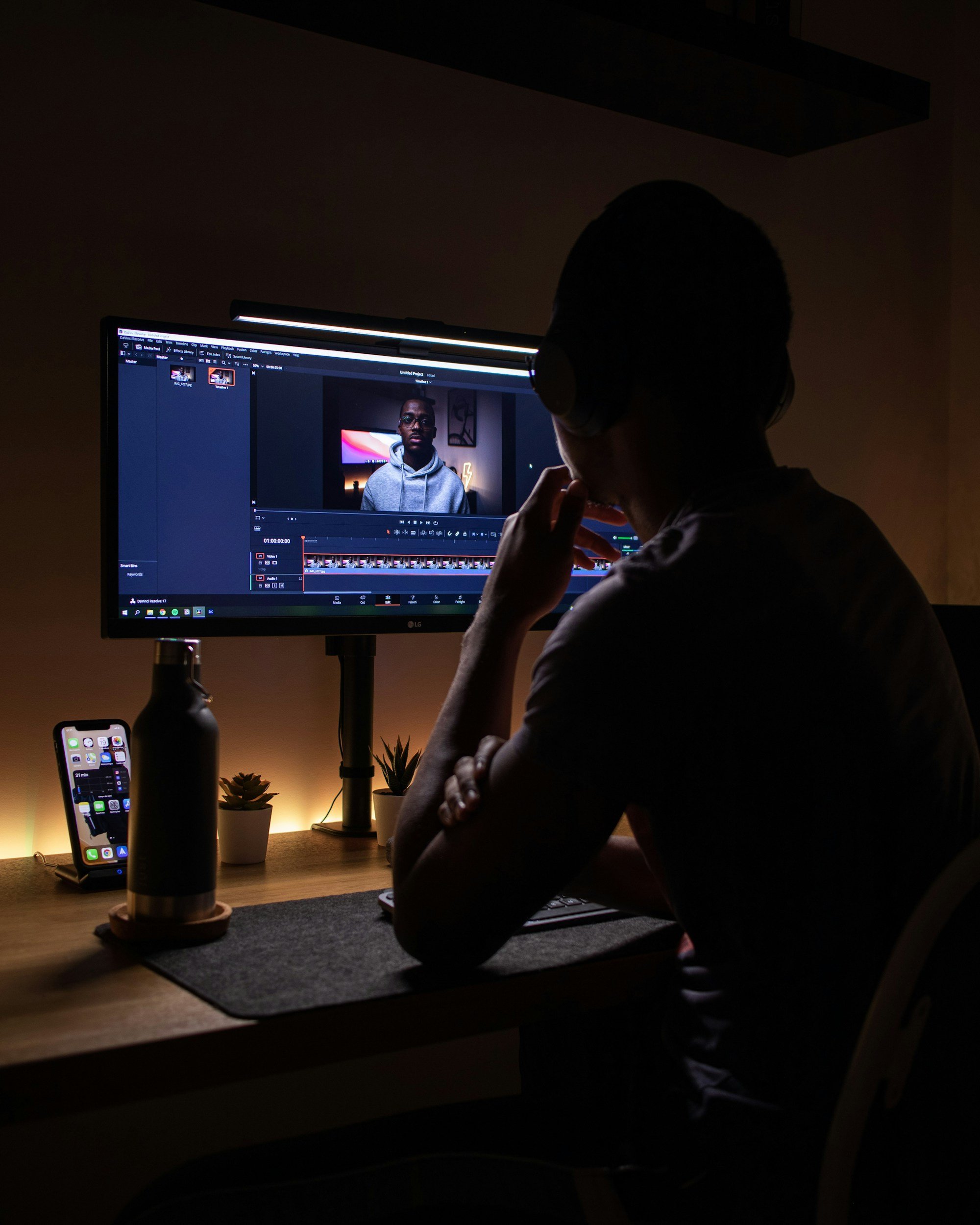 Person working on video editing on a computer in a dimly lit room, with a smartphone and plants on the desk.