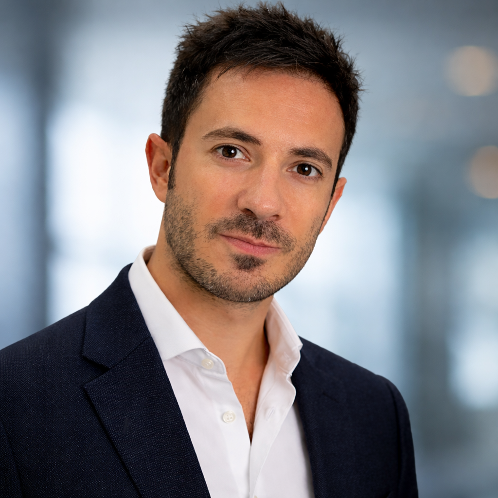 A professional man in a dark suit jacket and white collared shirt, with short dark hair and light facial hair, posing in front of a blurred office background.