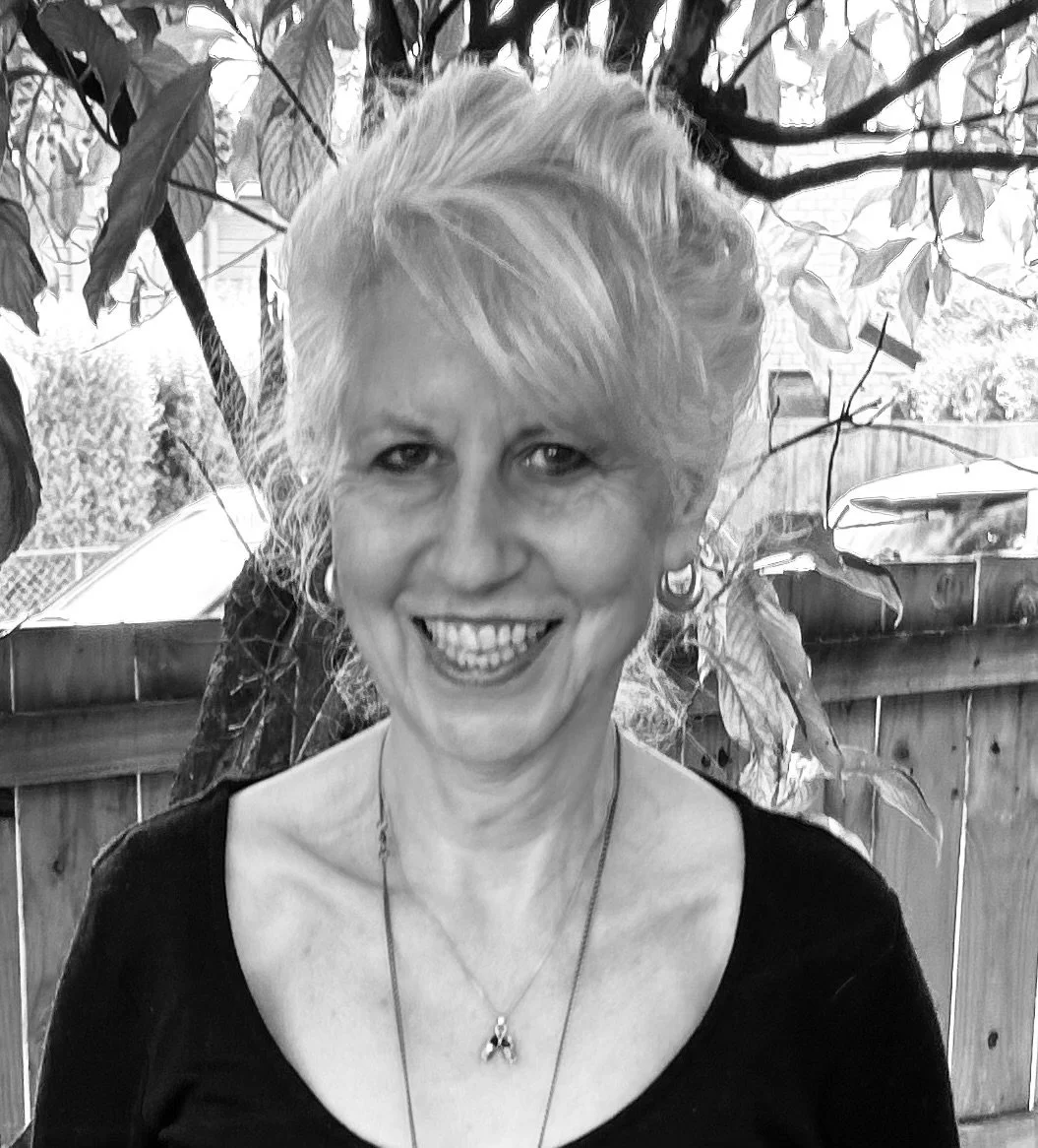 Black and white photo of a smiling middle-aged woman with short, light-colored hair, wearing earrings and necklaces, standing outdoors in front of a wooden fence and leafy branches.