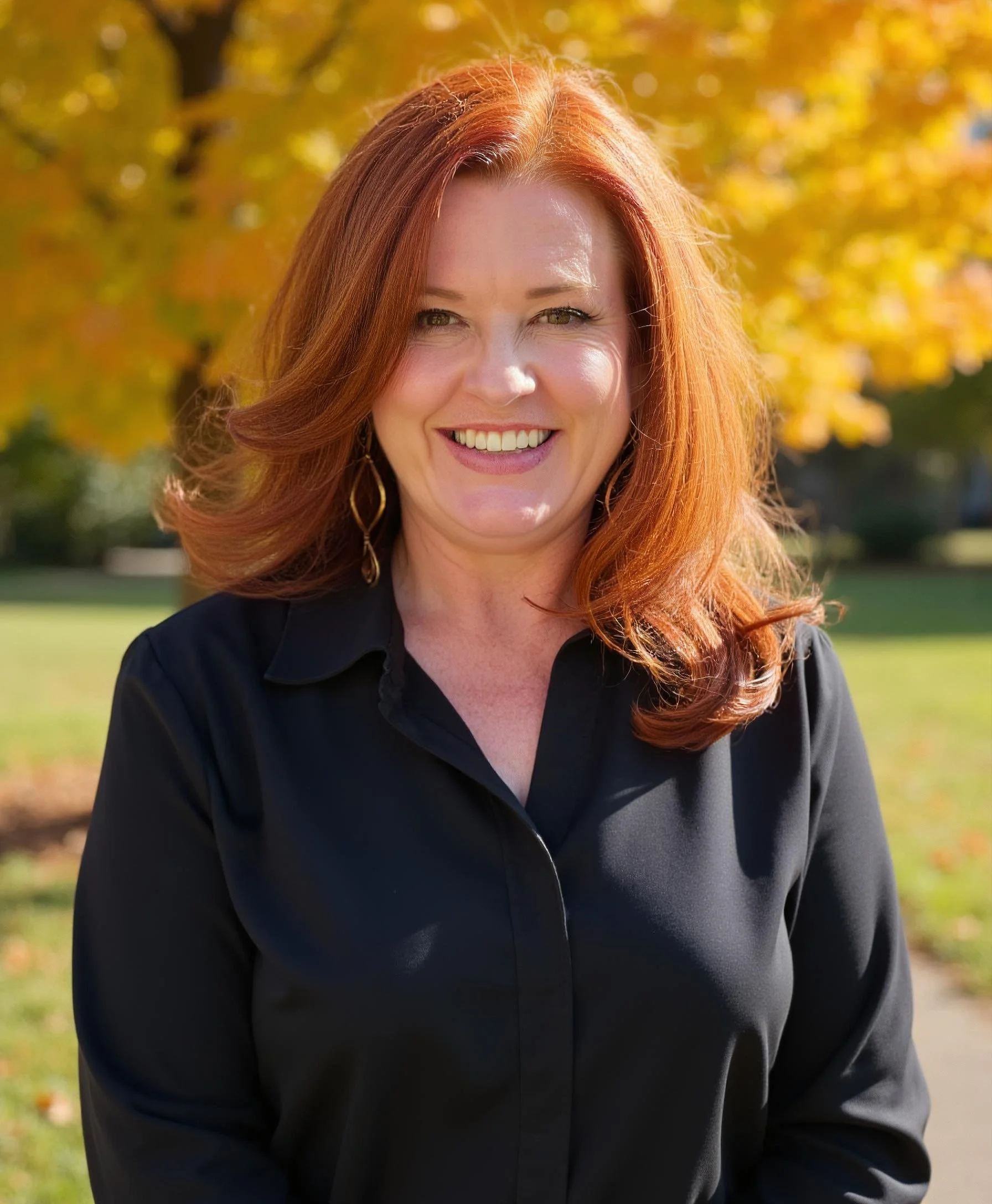 A smiling woman with red hair, wearing a black jacket, outdoors in fall with yellow and orange leaves in the background.