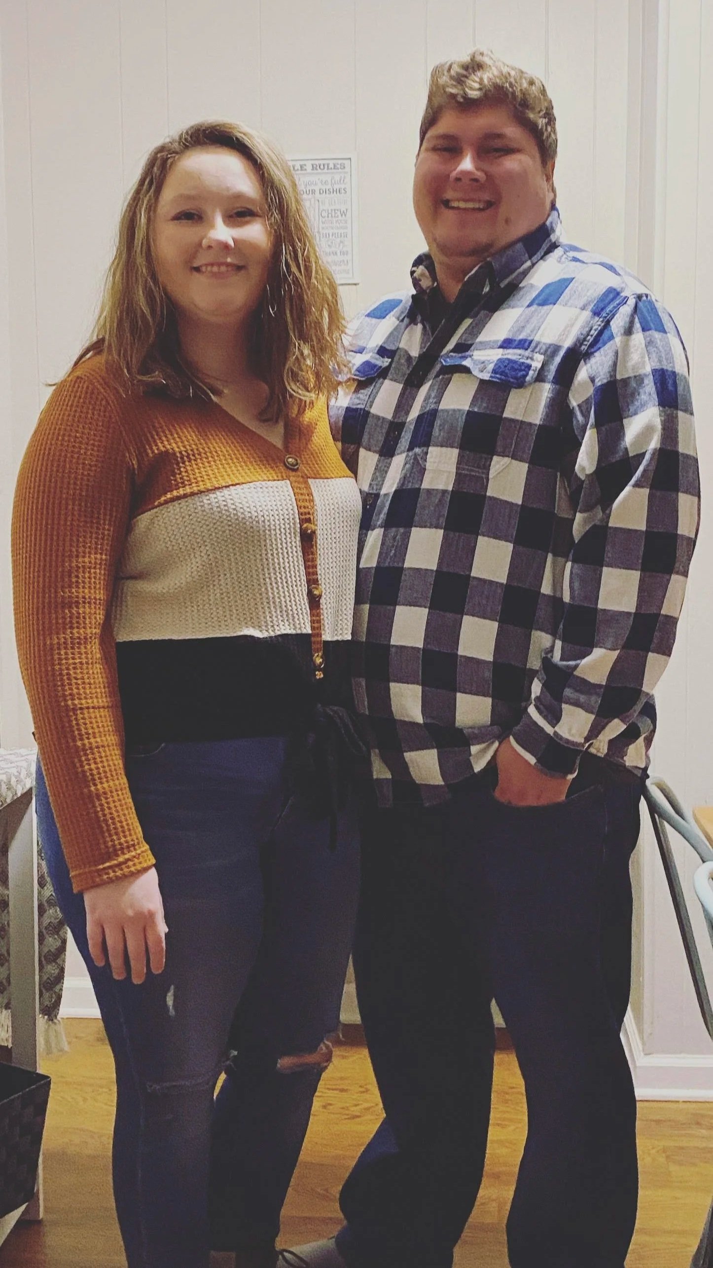A woman and a man standing close together indoors, smiling at the camera. The woman wears a multicolored button-up sweater and dark pants, and the man wears a plaid shirt and dark pants.