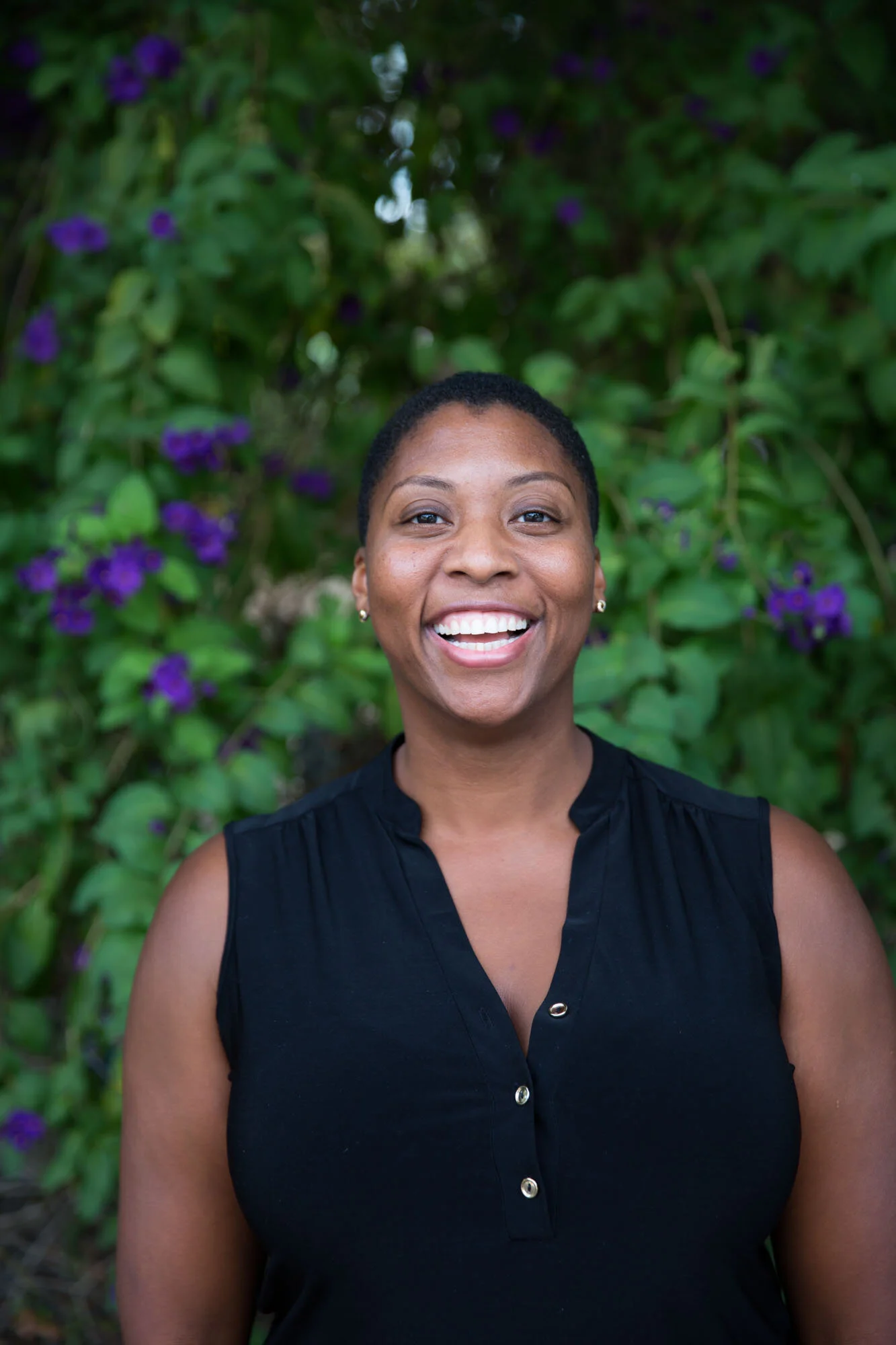 A smiling Black woman with short hair, wearing a sleeveless black top with buttons, standing outdoors in front of greenery with purple flowers.