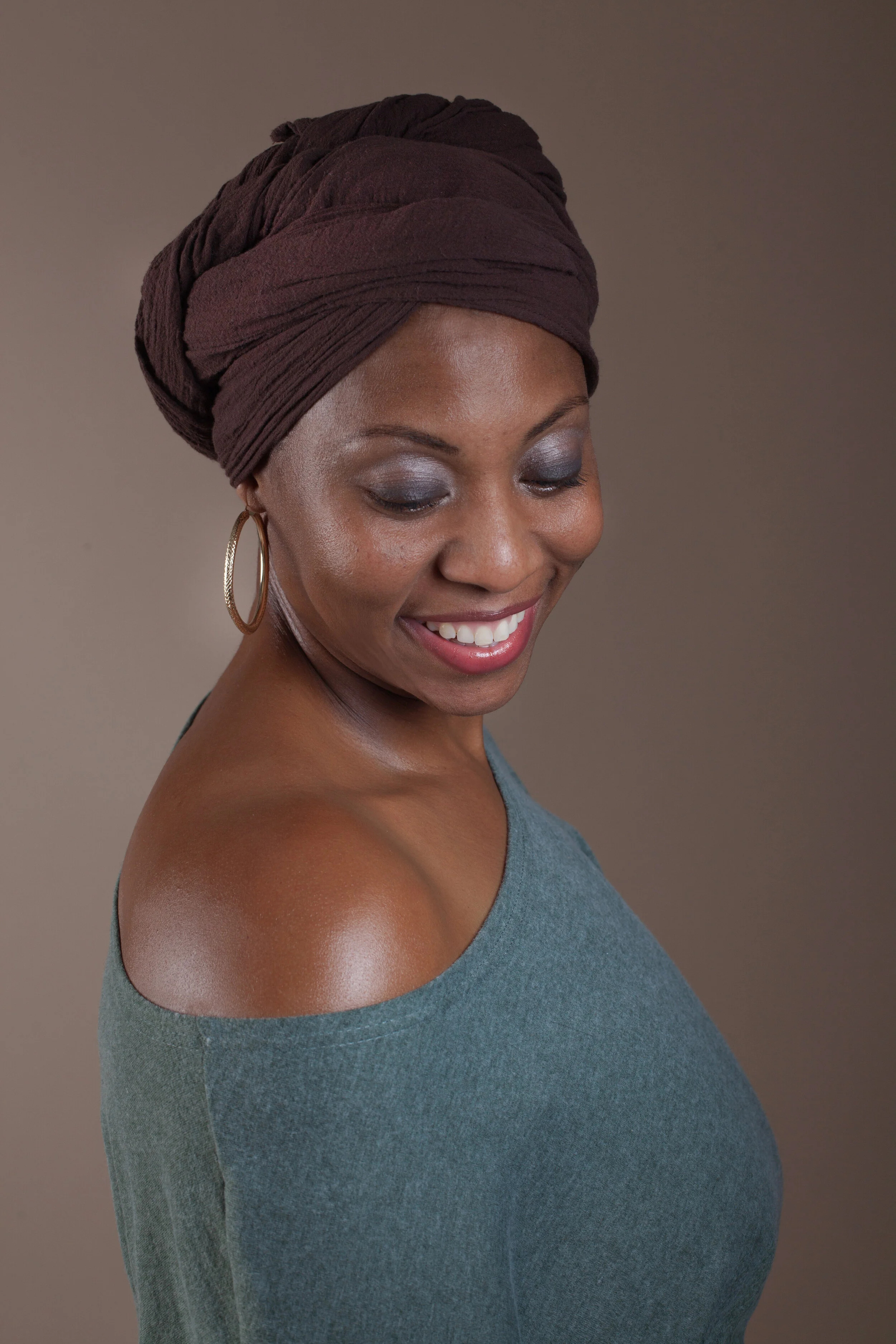 Melanie Larkins, founder of Kindred & Core, looking downward with a gentle smile, wearing a rich brown fabric headwrap and gold hoop earrings, showcasing hydrated, glowing skin.