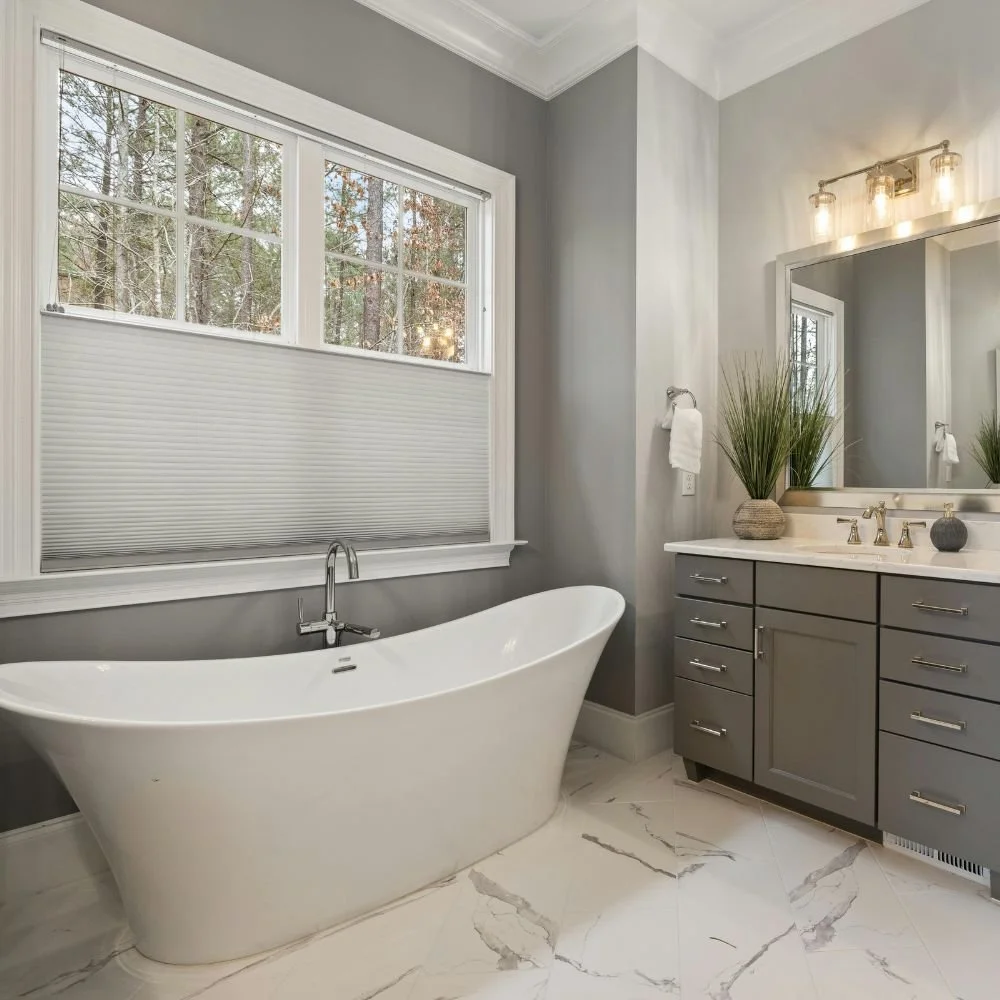 Modern bathroom with a freestanding bathtub beneath a large window and grey cabinetry with a white countertop and gold fixtures.