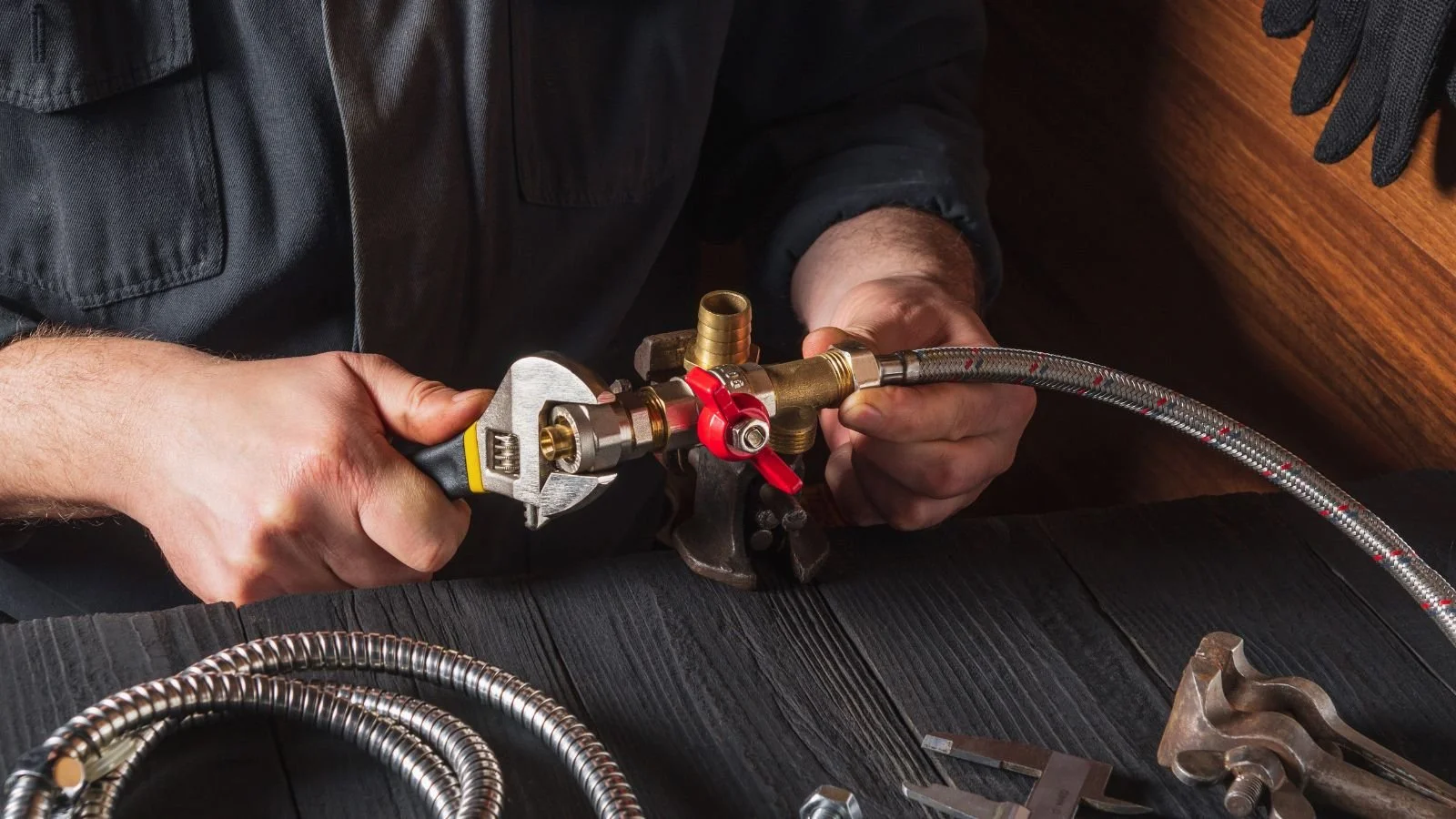 A person installing a flexible metal water hose onto a brass valve with a red handle, using a wrench, on a dark wooden surface.