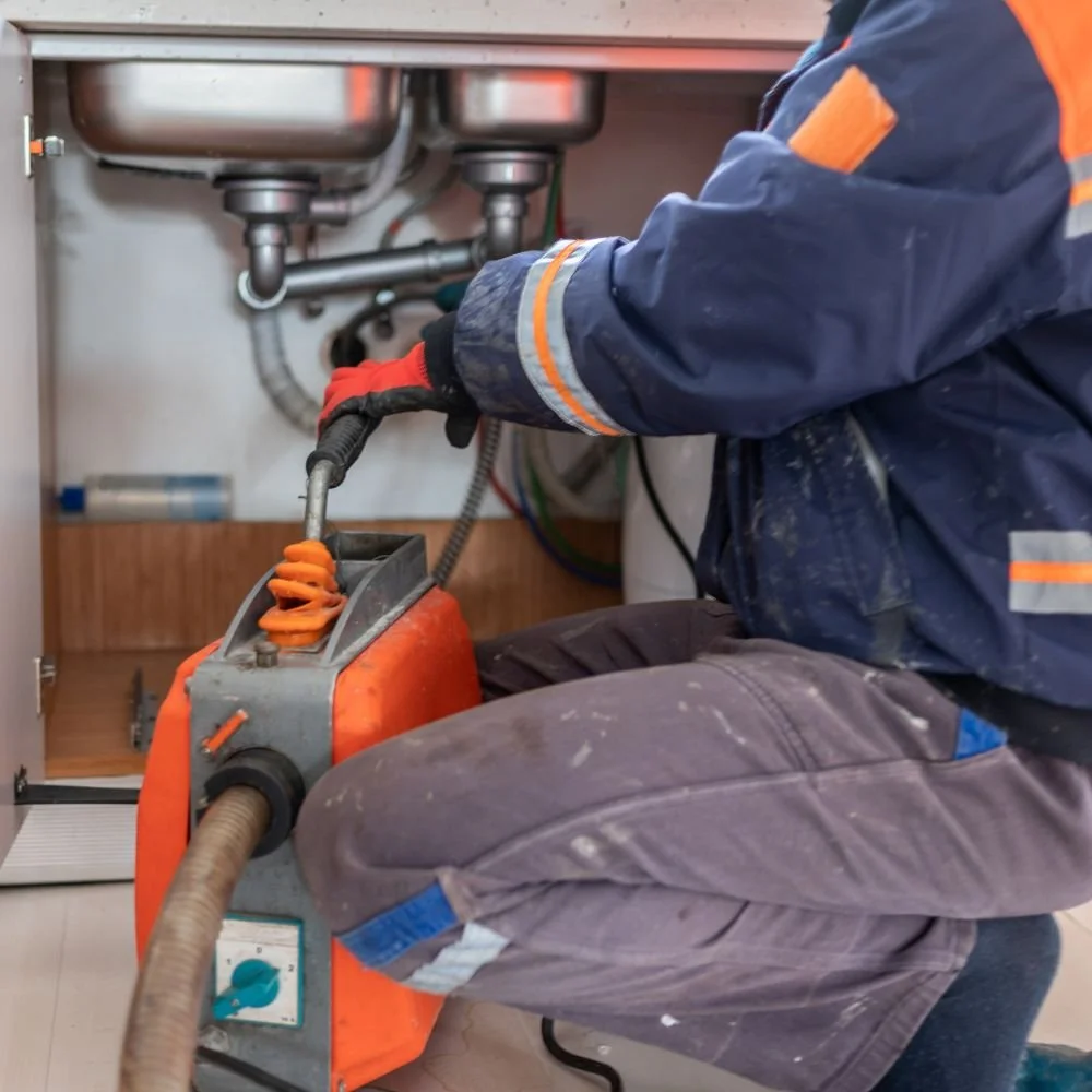 A technician working under a kitchen sink using a power drain cleaner to clear a clog.