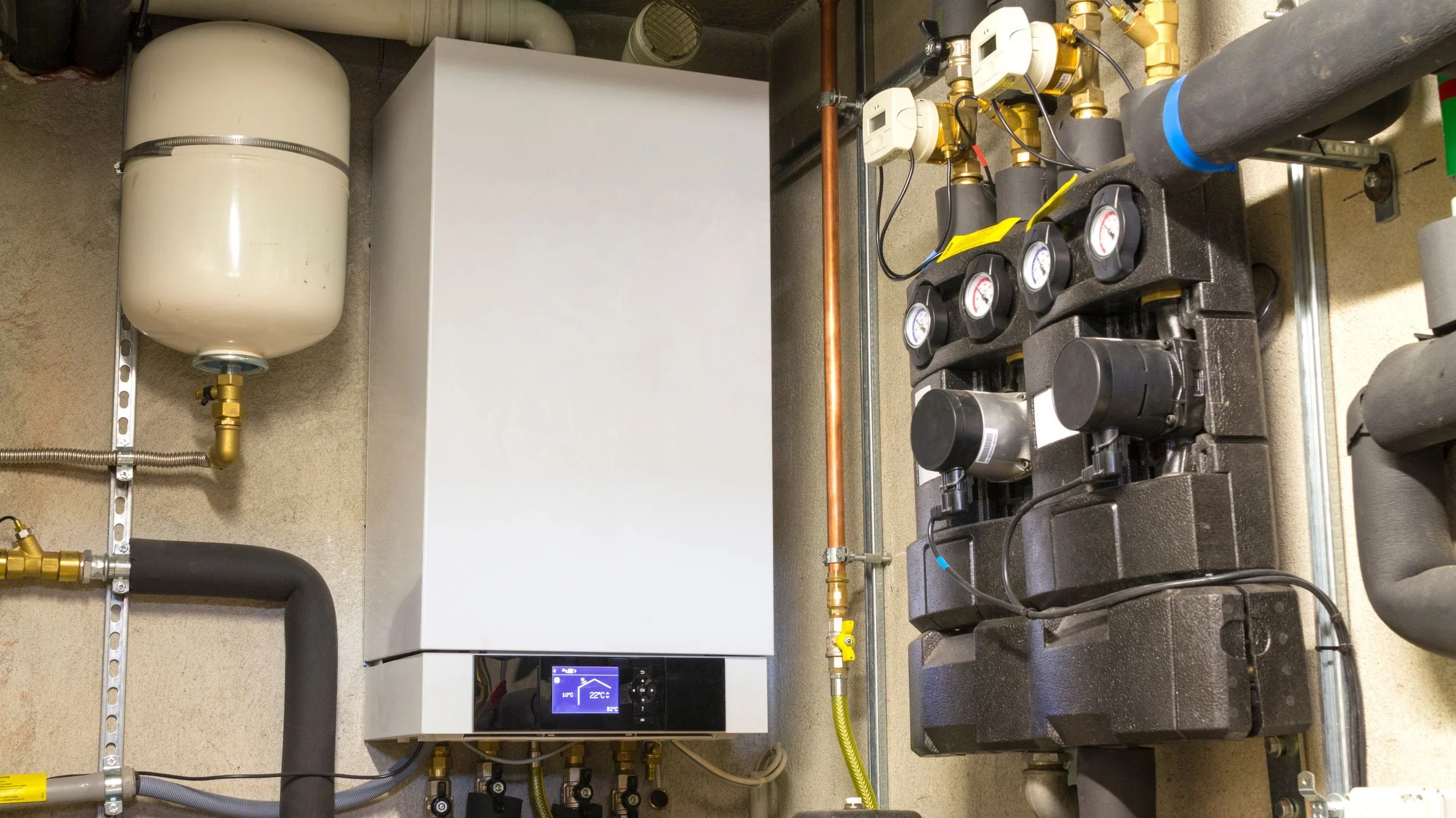 A mechanical room with a white tank, a digital control panel, and various pipes, valves, and gauges.